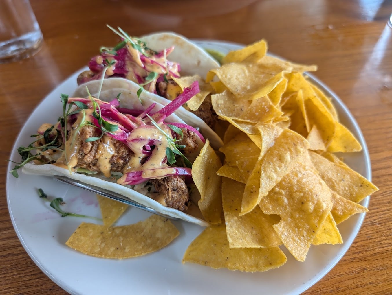 Two soft shell tacos filled with grilled chicken, pickled onions, greens, and sauce are served with a side of yellow tortilla chips on a white plate.