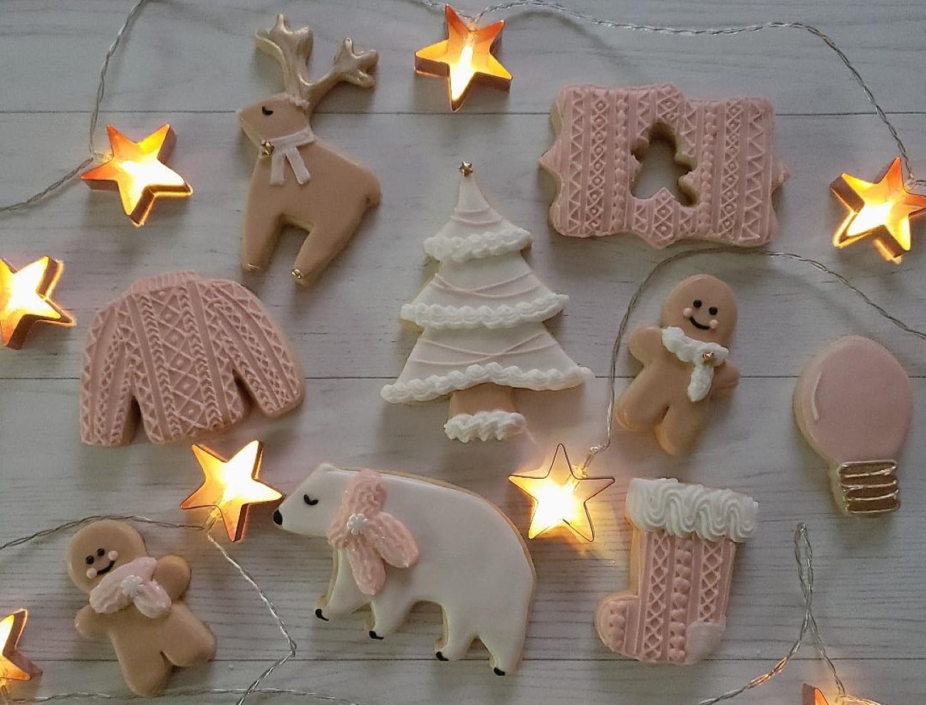 Seasonal cookies decorated in charming shapes such as a sweater, reindeer, tree, polar bear, mittens, stocking, and light bulb are artfully arranged on a white wooden surface and surrounded by glowing star string lights.