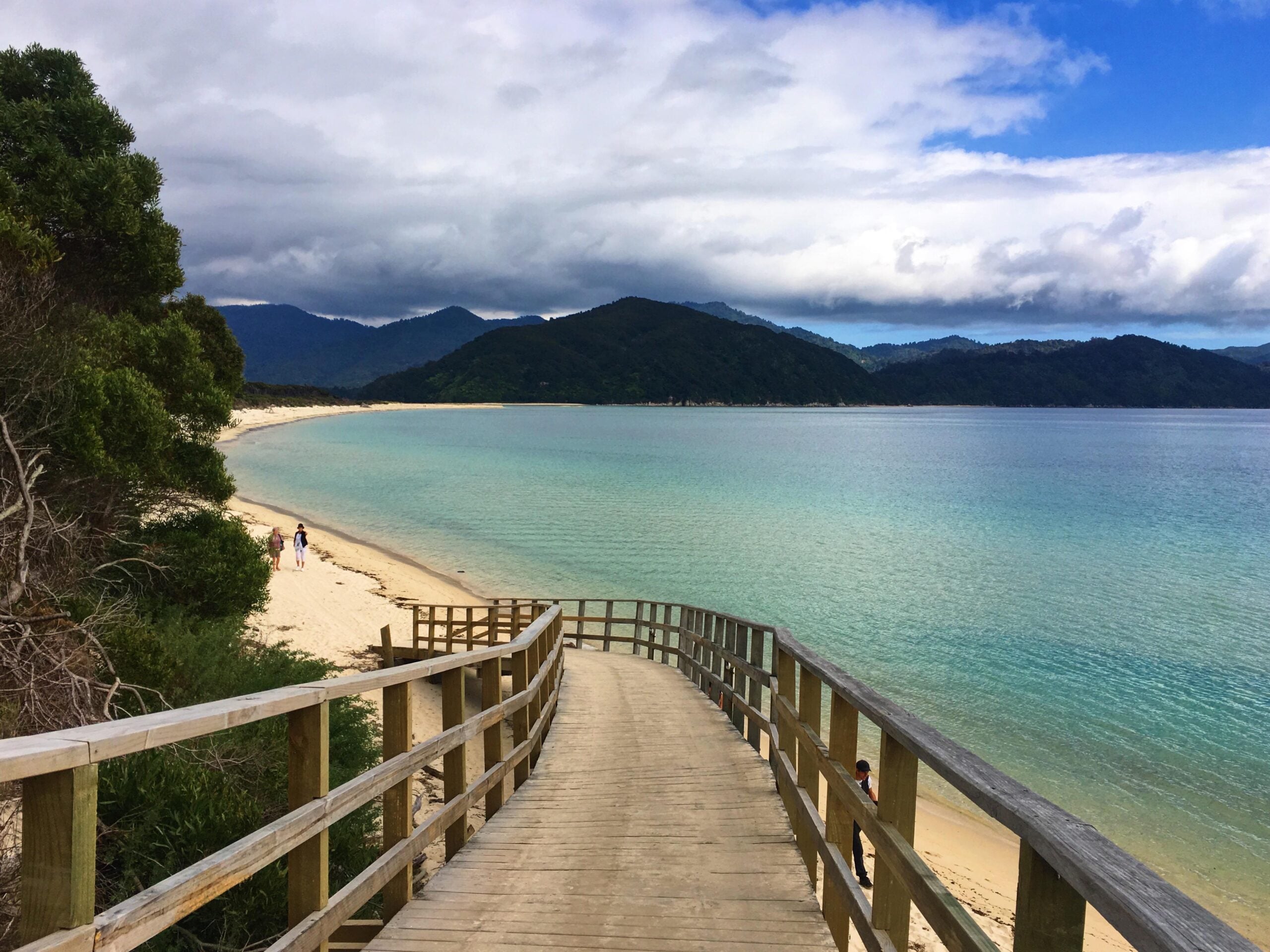 A wooden boardwalk leads to a sandy beach with turquoise water. Green hills and mountains are in the distance under a cloudy sky. A few people walk along the shore.