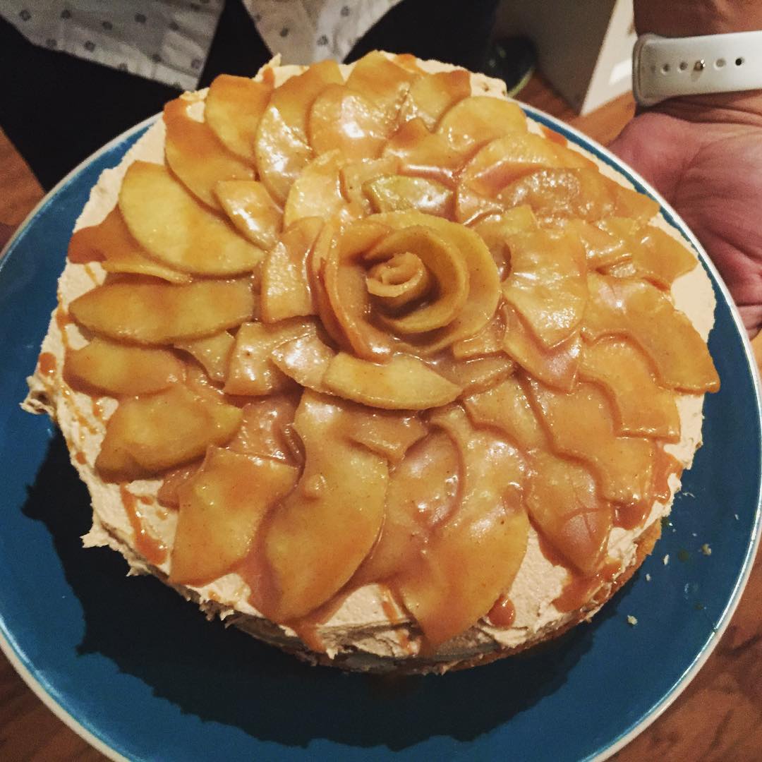 A round cake topped with thinly sliced caramelized apples arranged in a flower pattern, served on a blue plate. Someone is holding the plate with one hand, wearing a white watch.
