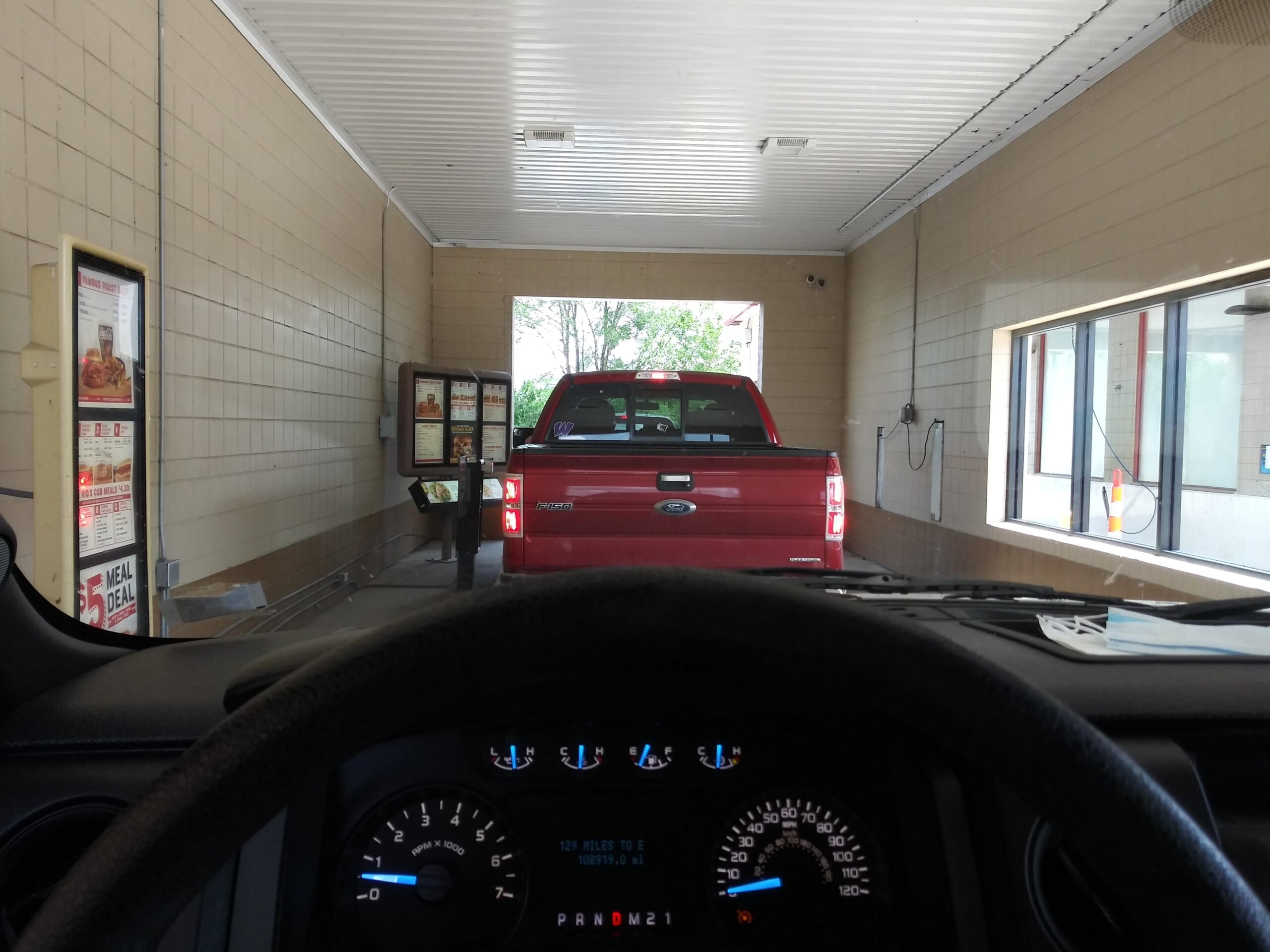 View from inside a vehicle at a drive-thru, with a red pickup truck in front, menu boards on the left, and a window on the right. Dashboard and steering wheel are visible in the foreground.