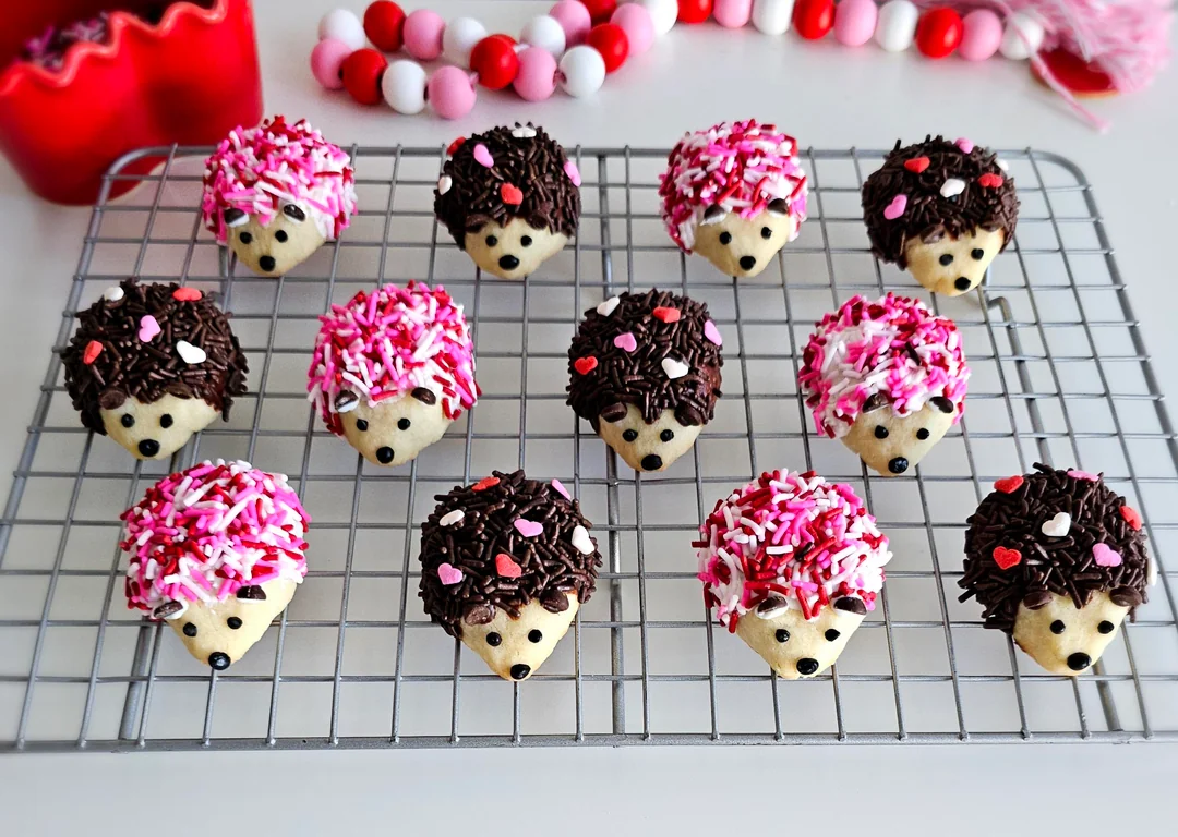 Decadent Desserts come to life with these adorable hedgehog-shaped cookies, topped with chocolate and pink sprinkles for fur and heart-shaped decorations, all beautifully displayed on a cooling rack with red and white bead garlands in the background.