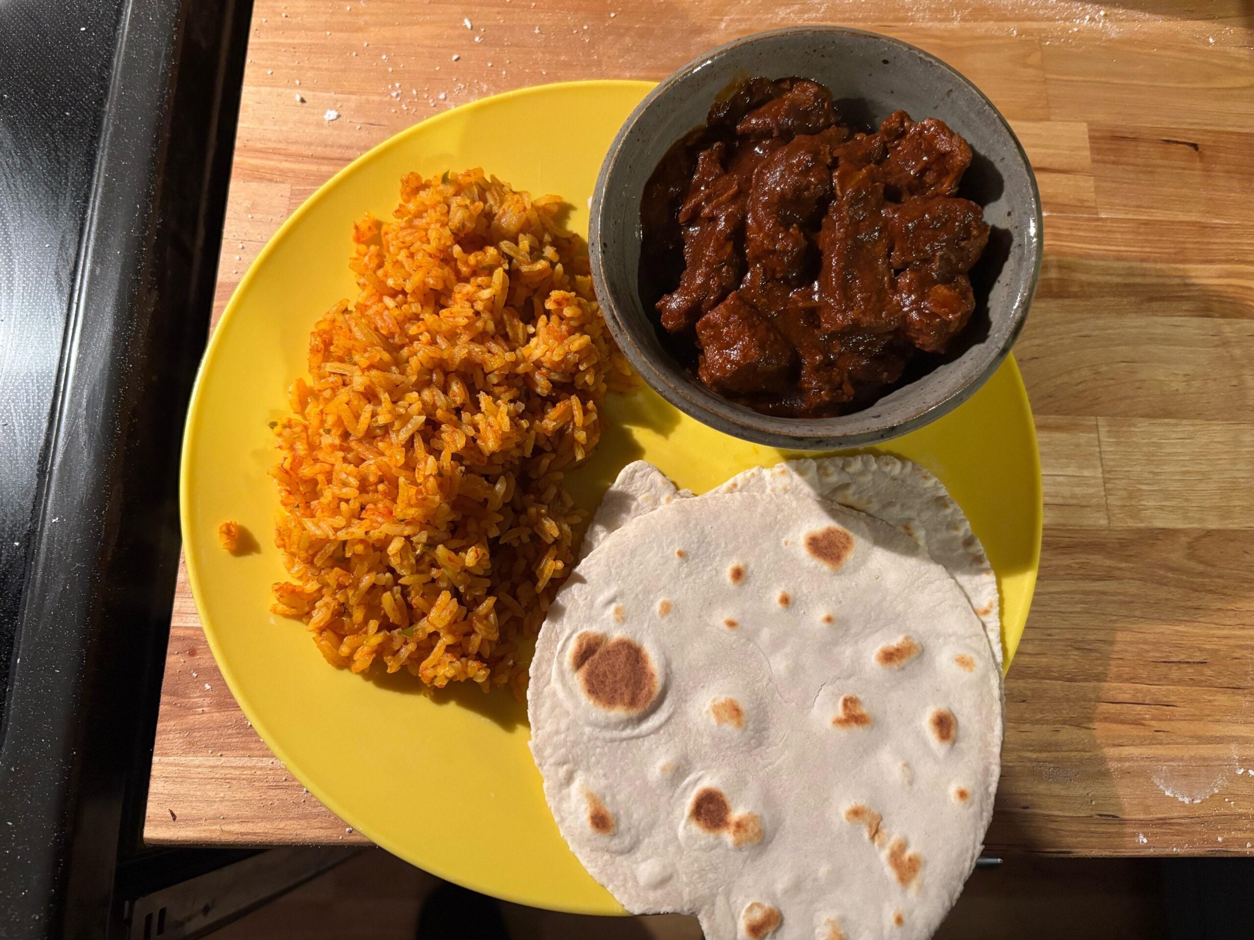 A yellow plate with orange rice, two pieces of flatbread, and a small bowl of dark, saucy meat stew, set on a wooden surface.