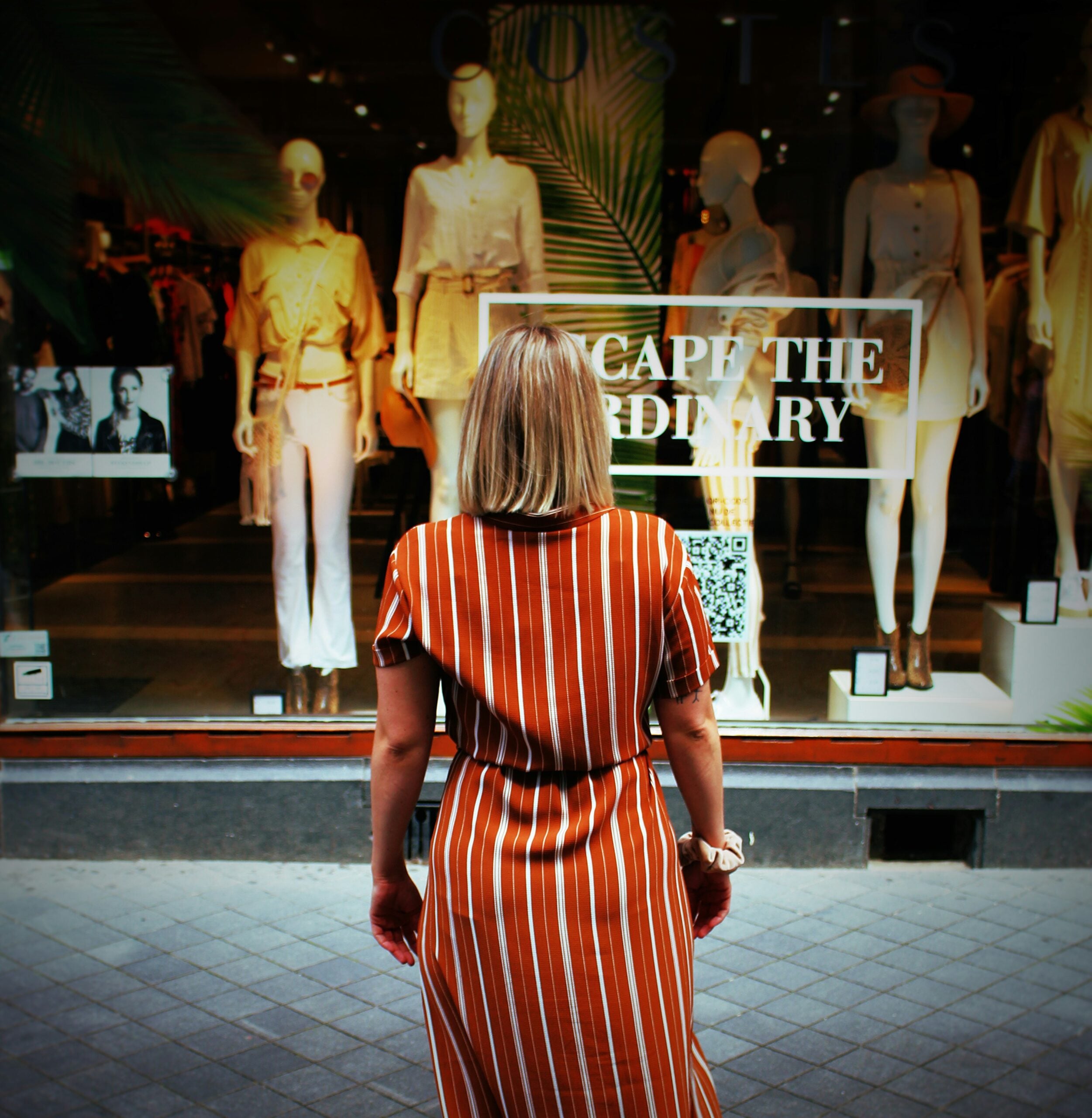 A woman in a striped dress stands facing a store window displaying mannequins and a sign that reads “ESCAPE THE ORDINARY.”