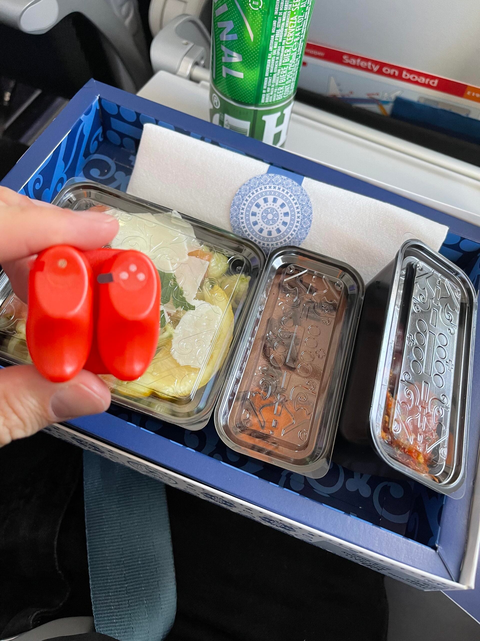 A hand holds red salt and pepper shakers over an airline meal tray with three covered dishes, a napkin, and a can of Perrier sparkling water on an airplane tray table.