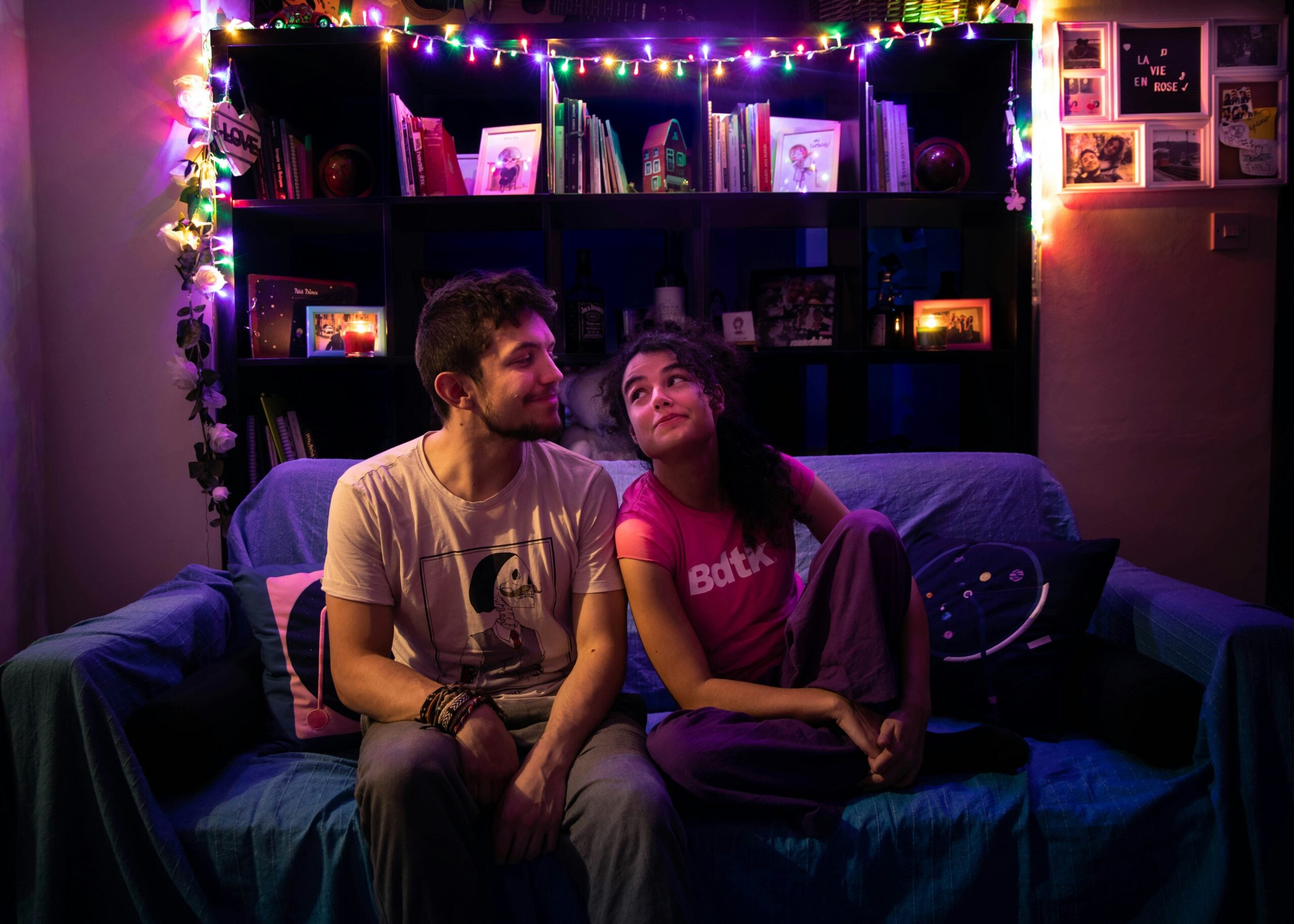 A man and woman sit on a blue couch in a cozy room with colorful string lights, shelves of books, and framed photos in the background. They smile and look at each other warmly.