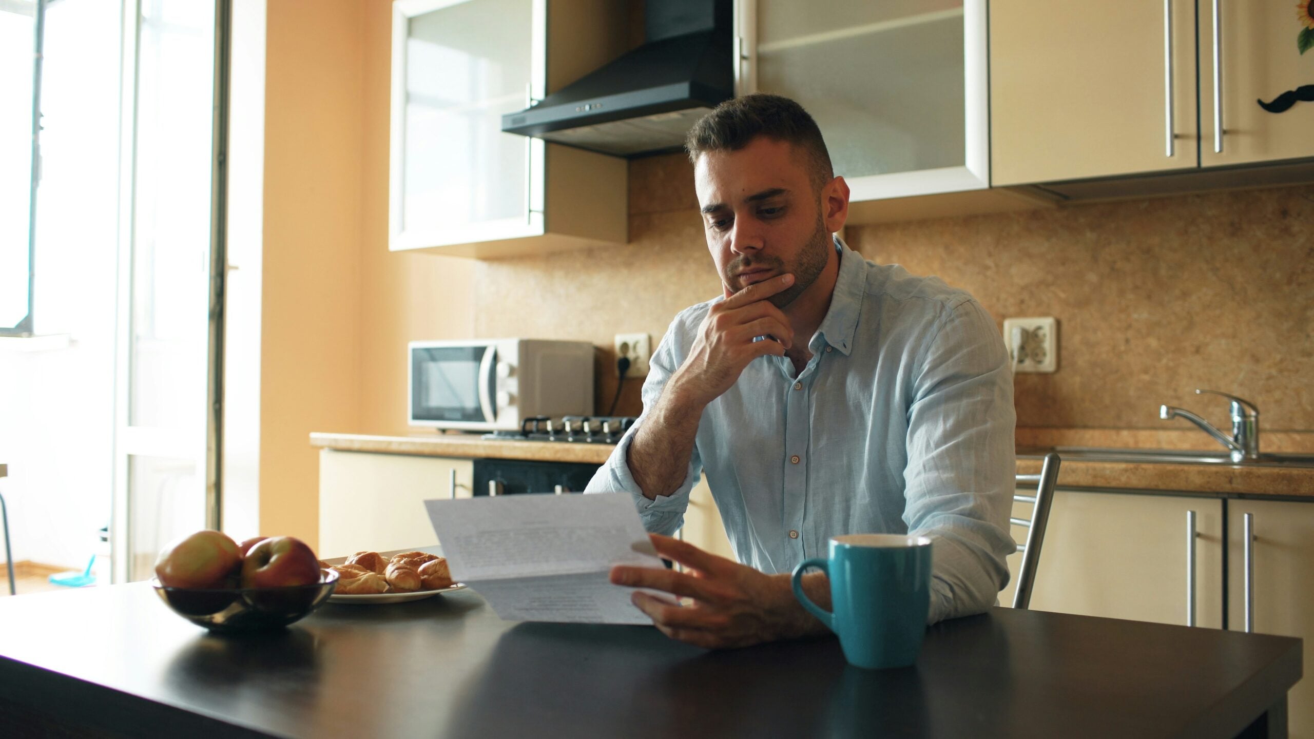 A man sits at a kitchen table, looking thoughtfully at a piece of paper. There is a blue mug, a fruit bowl, and a plate with pastries on the table. The kitchen is tidy and well-lit.