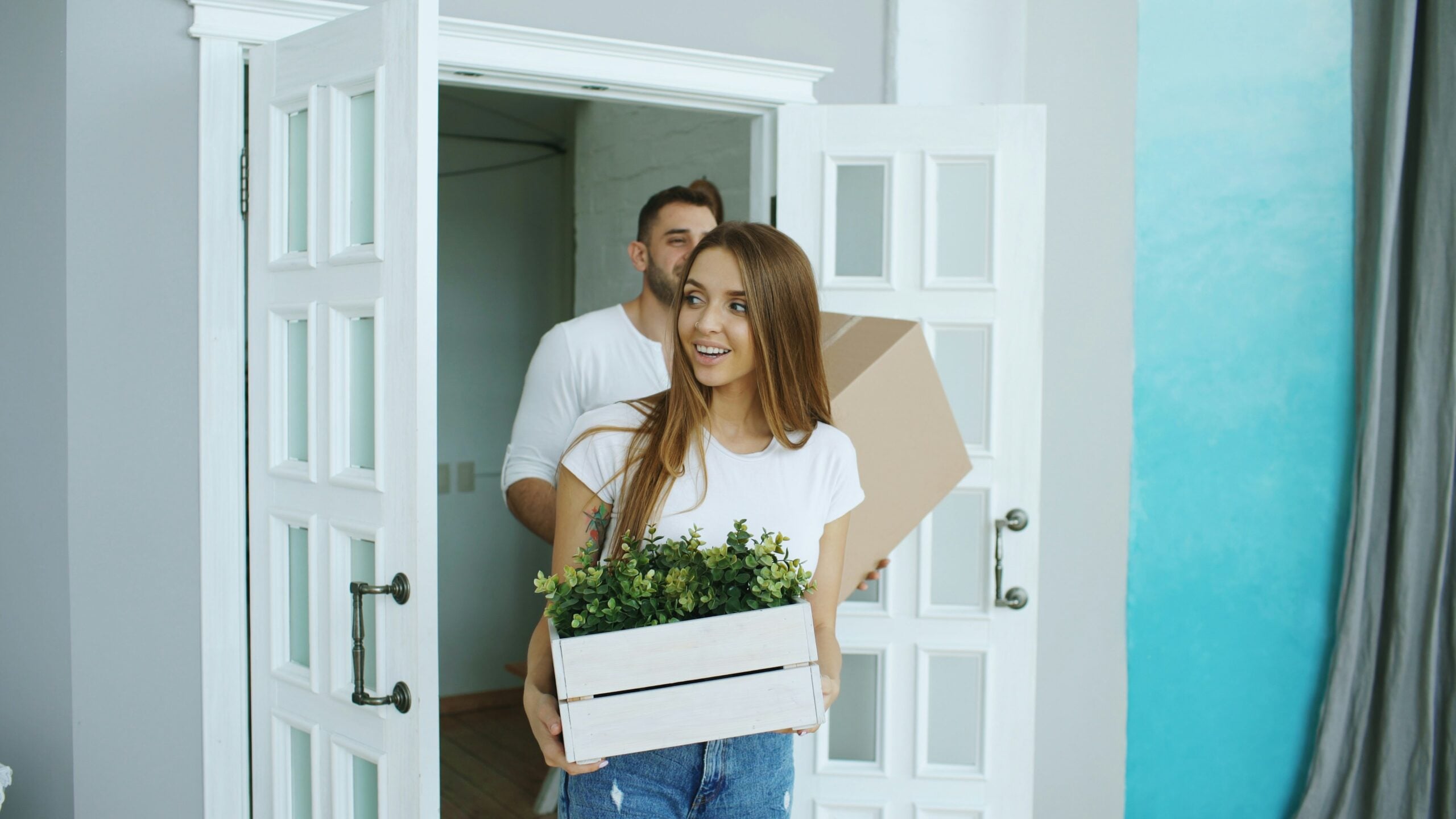 A woman carrying a box with a potted plant enters a bright room, followed by a man holding a cardboard box. Both are smiling and dressed casually.