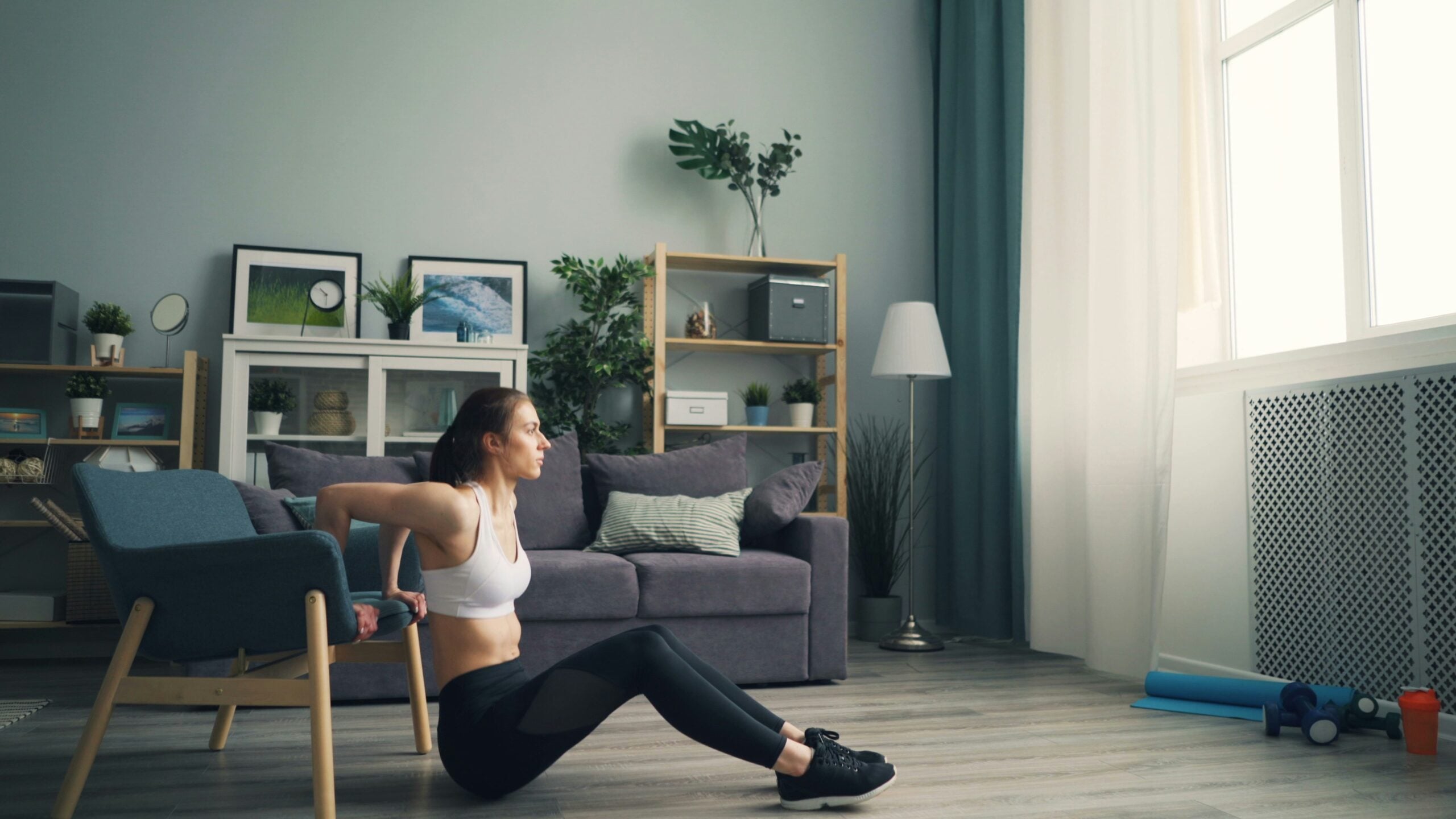 A woman in workout clothes does tricep dips using a chair in a living room with a sofa, plants, shelves, and framed pictures in the background. Natural light comes through a large window.