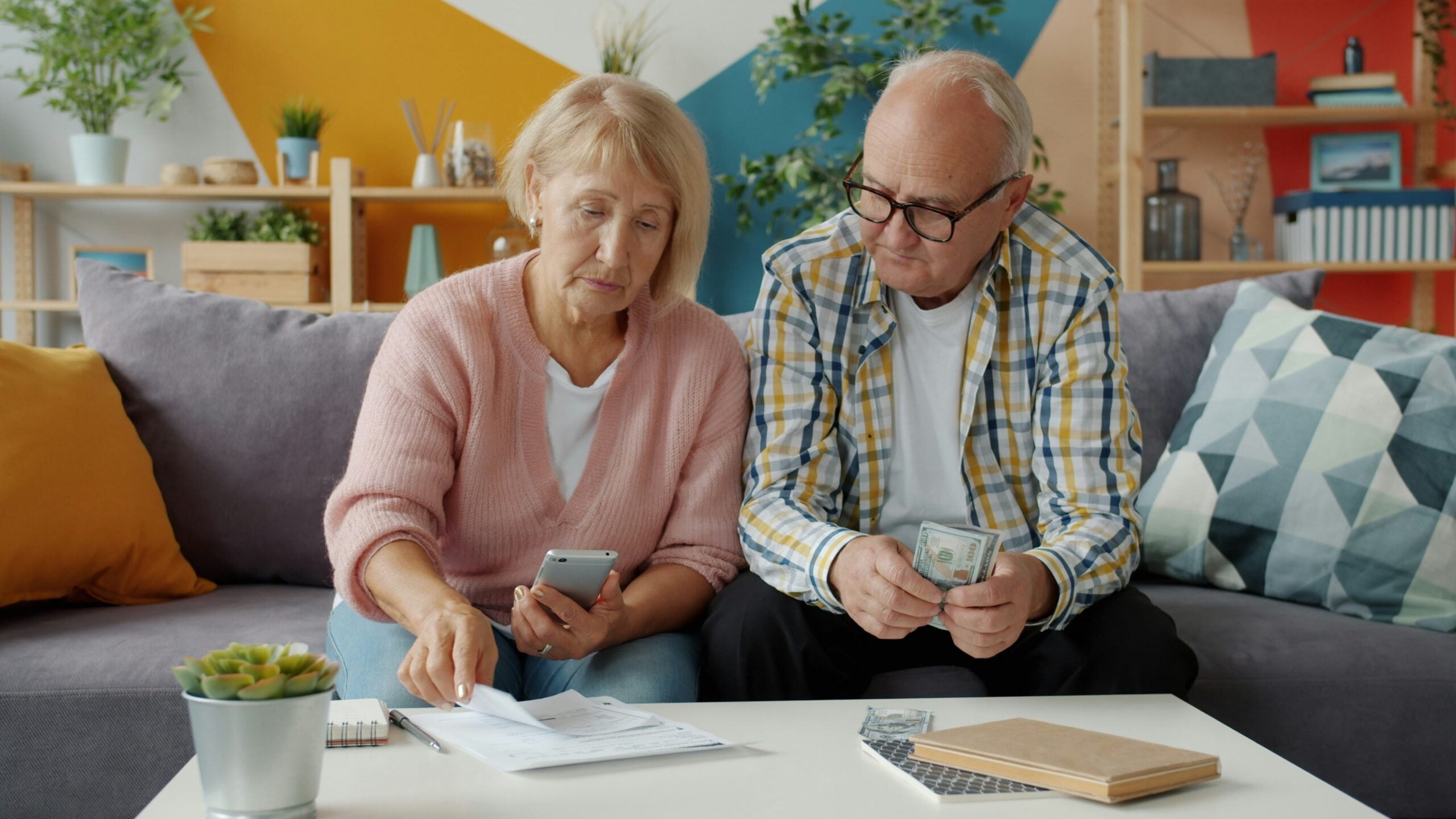 An older couple sits on a couch at a coffee table, reviewing papers and holding cash as they discuss their retirement savings. The woman holds a phone while pointing at documents, and the man counts money. The room is decorated with plants and colorful walls.