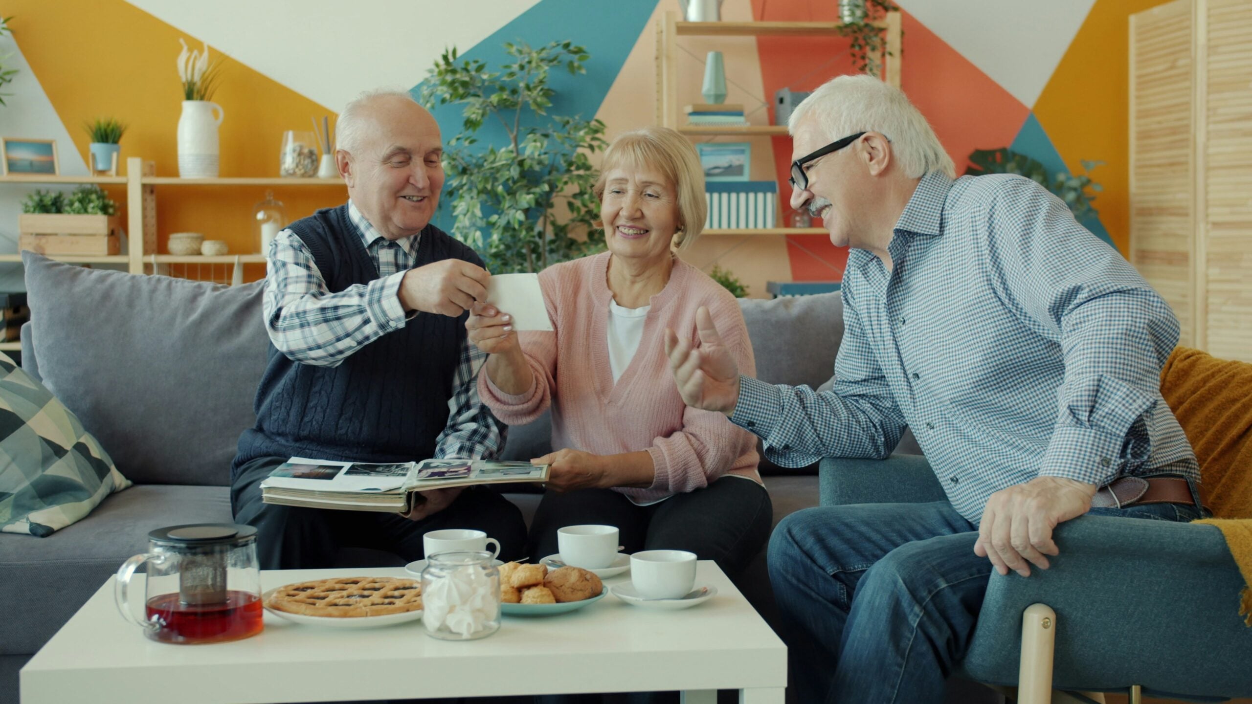 Three smiling older adults sit on a couch, sharing memories and snacks with coffee and pastries on the table in front of them, in a cozy, colorful living room—enjoying the comforts made possible by their thoughtful retirement savings.