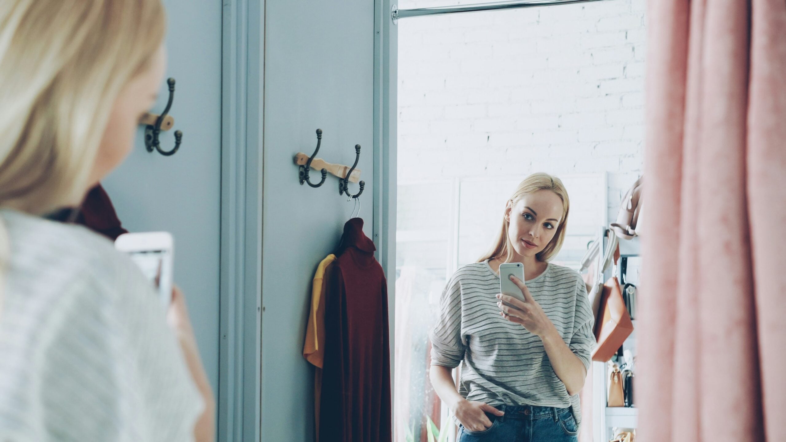 A woman stands in front of a mirror taking a selfie with her phone. She is wearing a striped shirt and jeans. Clothes and bags hang on the wall and a rack nearby. The room is bright with natural light.