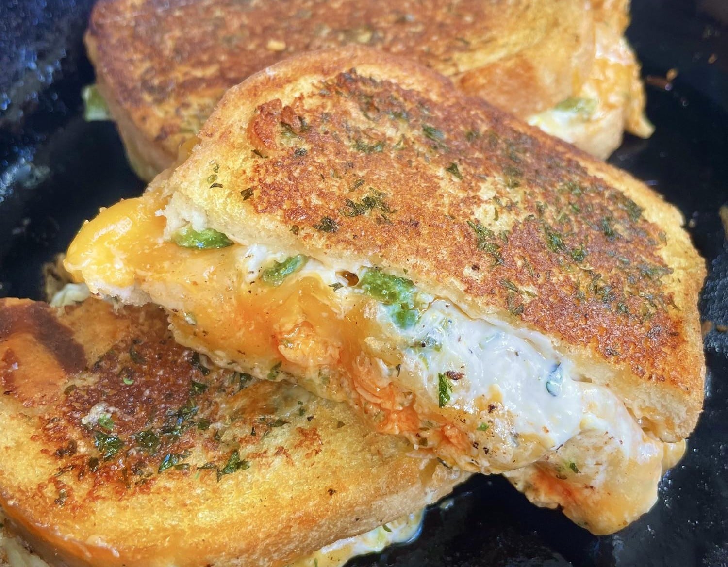 Close-up of one of the best grilled cheese sandwiches: a golden crust with melted cheese and herbs, cut in half to reveal a creamy, cheesy filling with green peppers, resting on a black surface.
