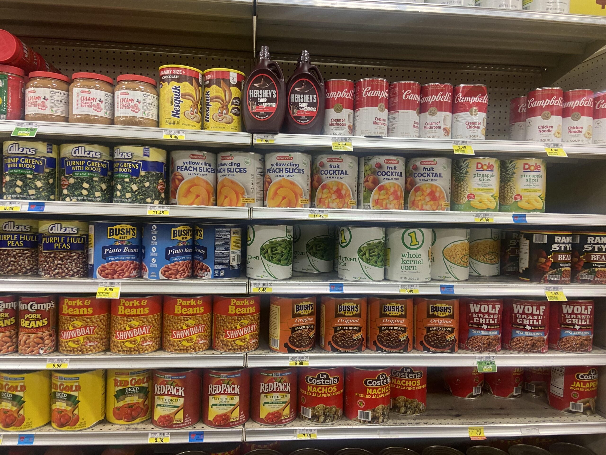 Grocery store shelves stocked with canned beans, vegetables, fruit, tomato products, and bottles of chocolate syrup, all neatly organized in rows.