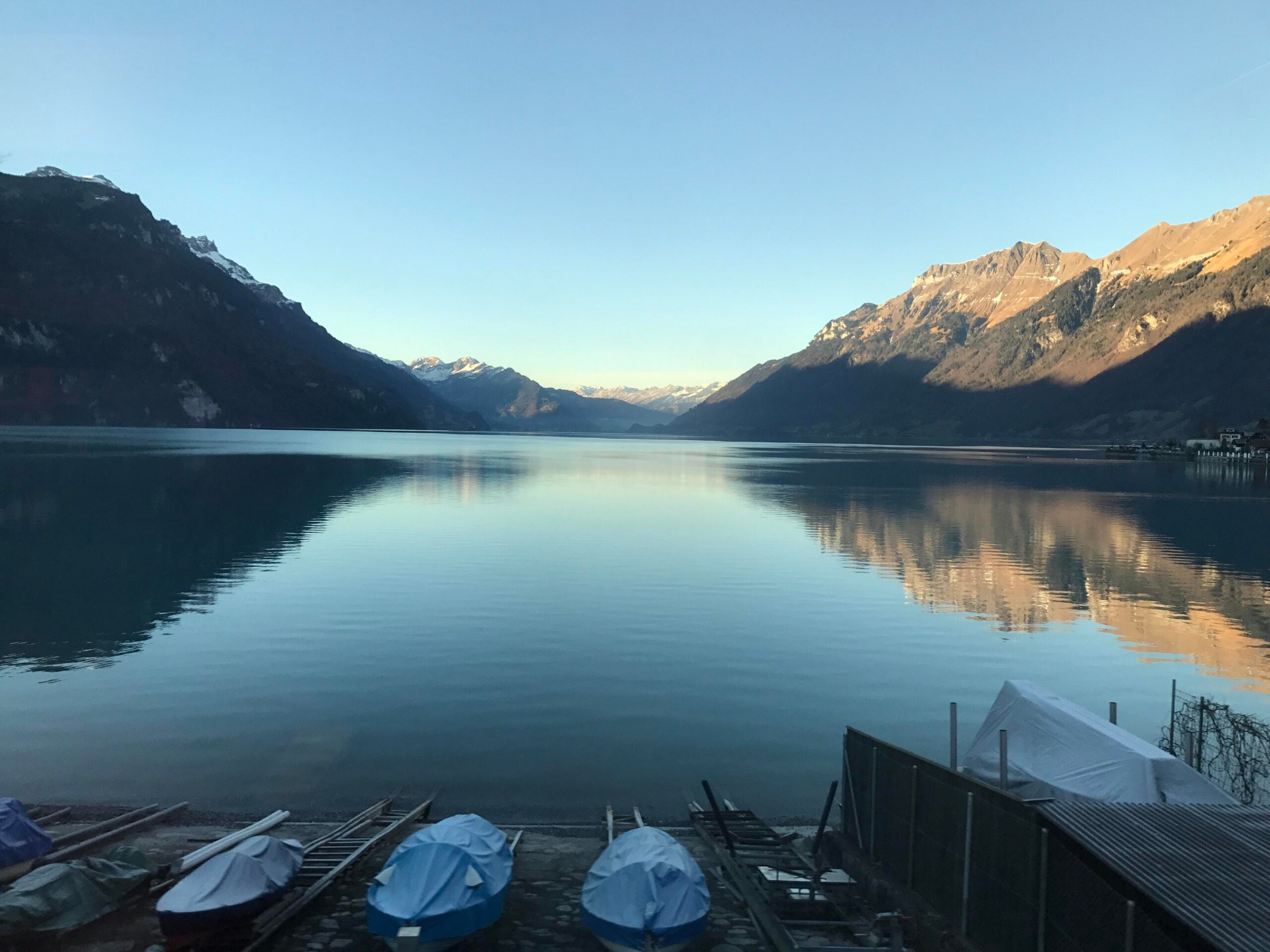 A calm lake reflects surrounding mountains at sunrise, with small boats covered on the shore in the foreground and clear blue skies above.