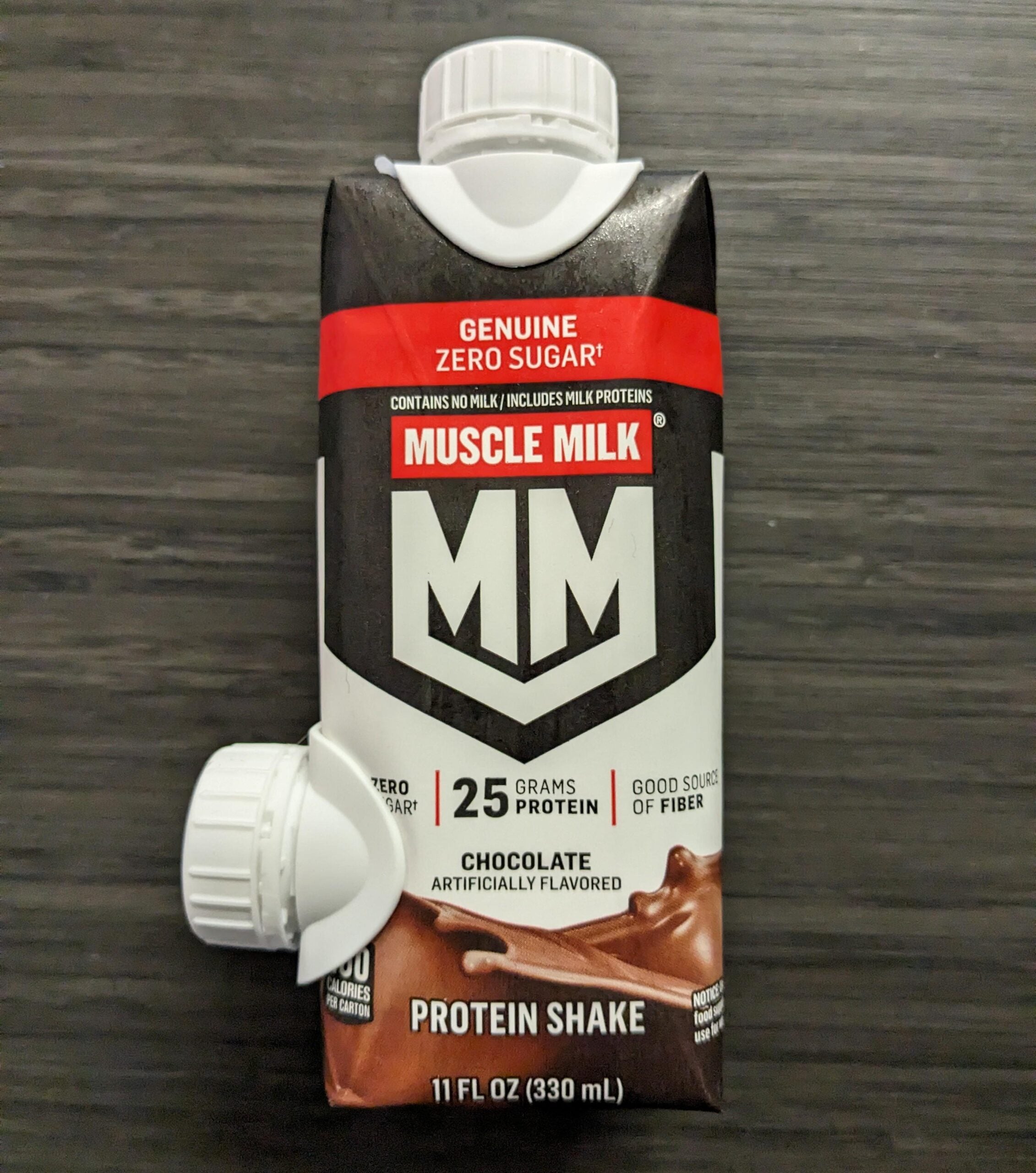 A carton of Muscle Milk chocolate protein shake with two white caps, labeled “25 grams protein” and “zero sugar,” sits on a dark wood surface. The carton is 11 fl oz (330 mL) and is chocolate artificially flavored.