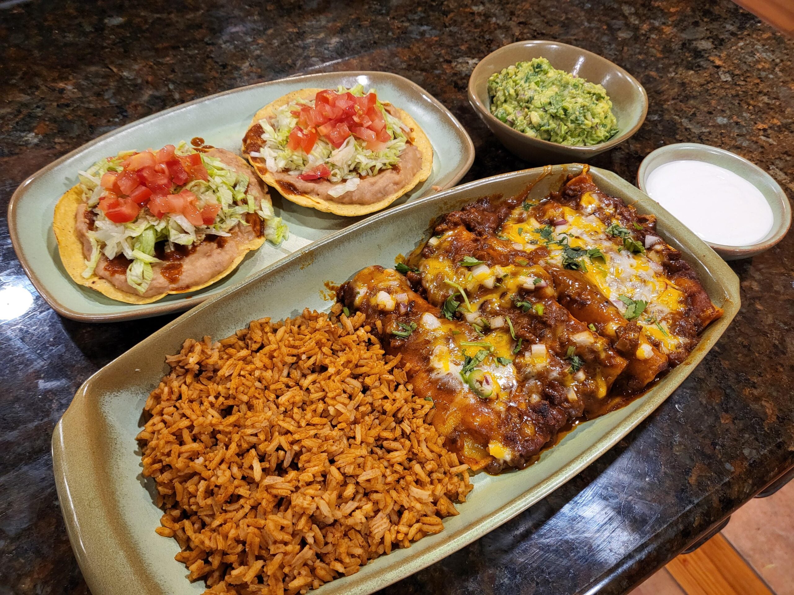 A plate with enchiladas topped with cheese and sauce, Mexican rice, two tostadas with refried beans, lettuce, tomatoes, a bowl of guacamole, and a small bowl of sour cream on a dark countertop.