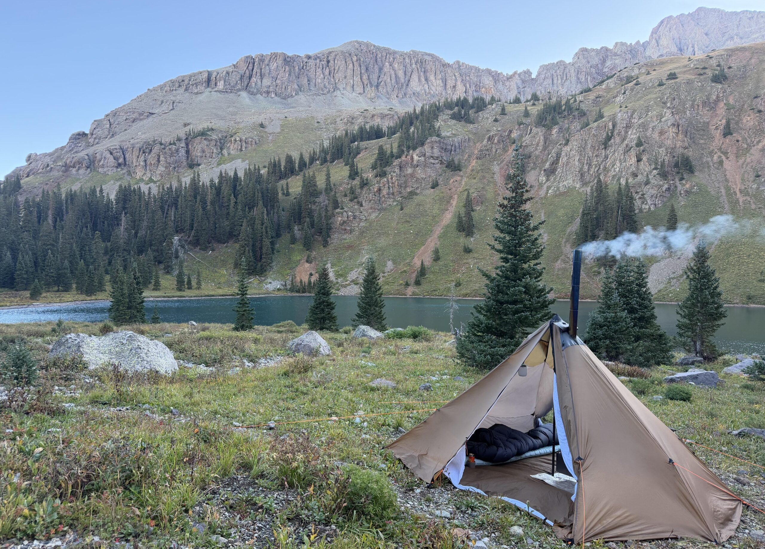 A brown tent with smoke coming from a stove pipe sits on grass by a lake, surrounded by pine trees and mountains under a clear sky.