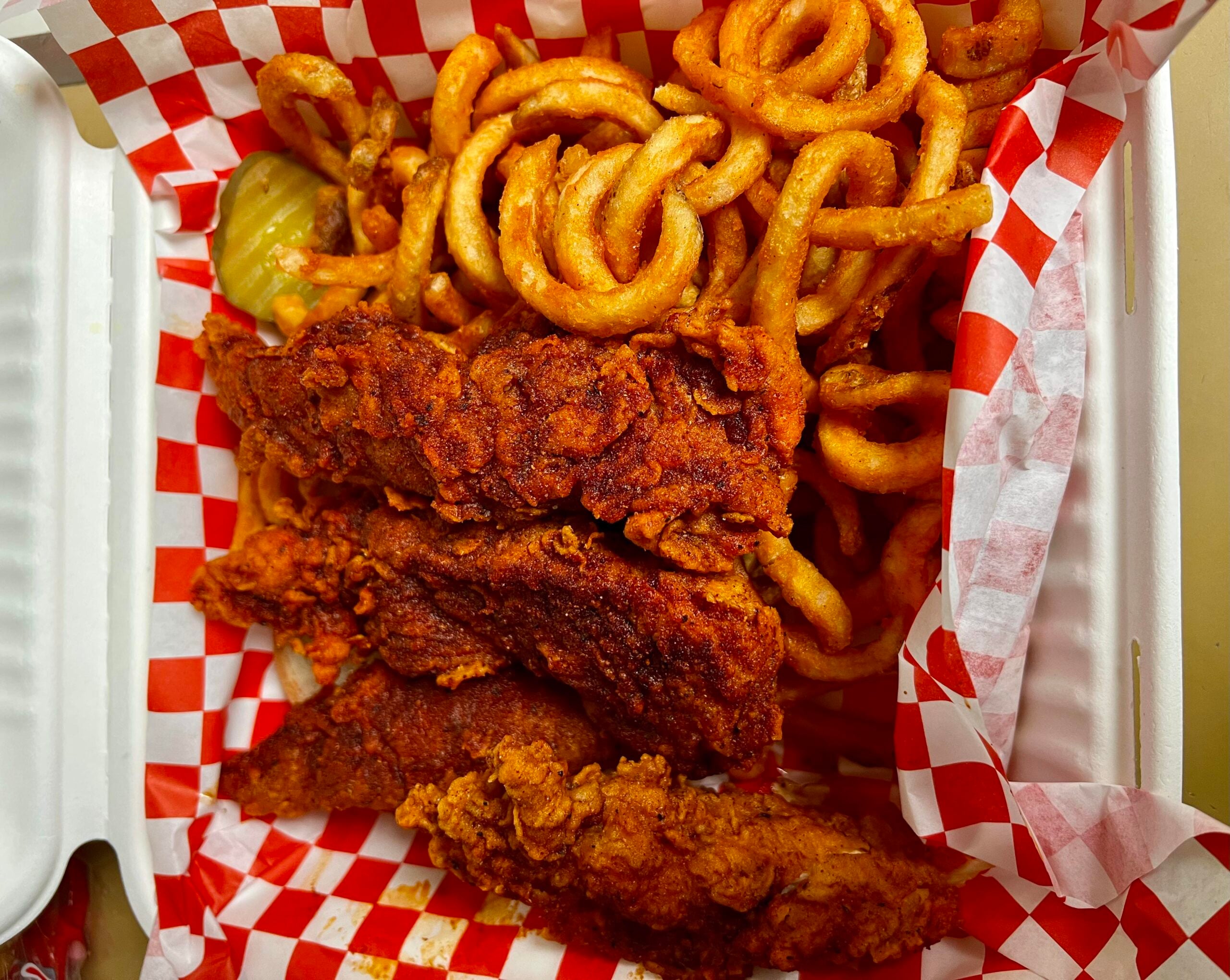 Three spicy fried chicken tenders and a pile of curly fries sit on red and white checkered paper in a takeout box, with a few pickle slices visible on the side.
