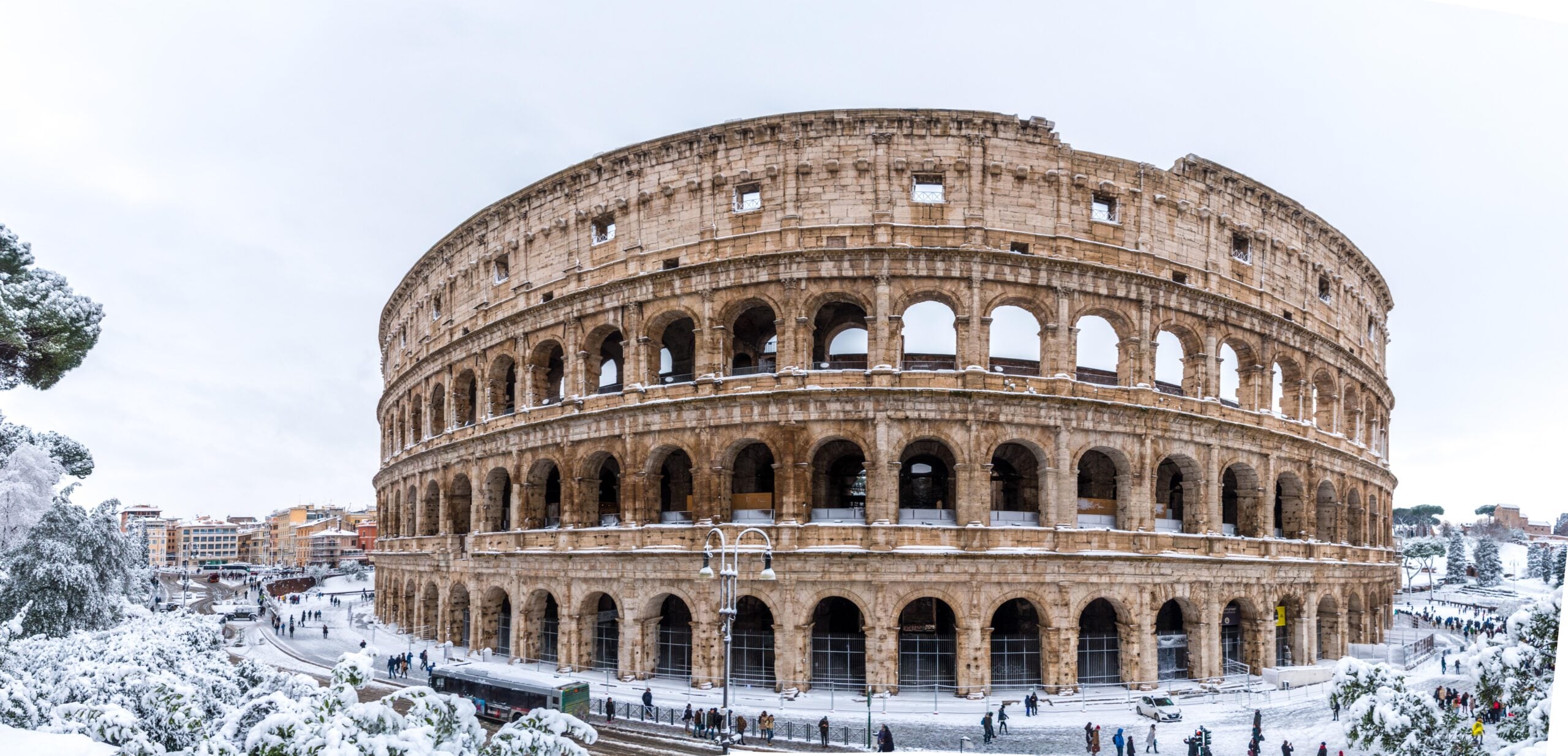 The Colosseum in Rome covered with snow; people walk nearby while trees and buildings around are also blanketed in snow.
