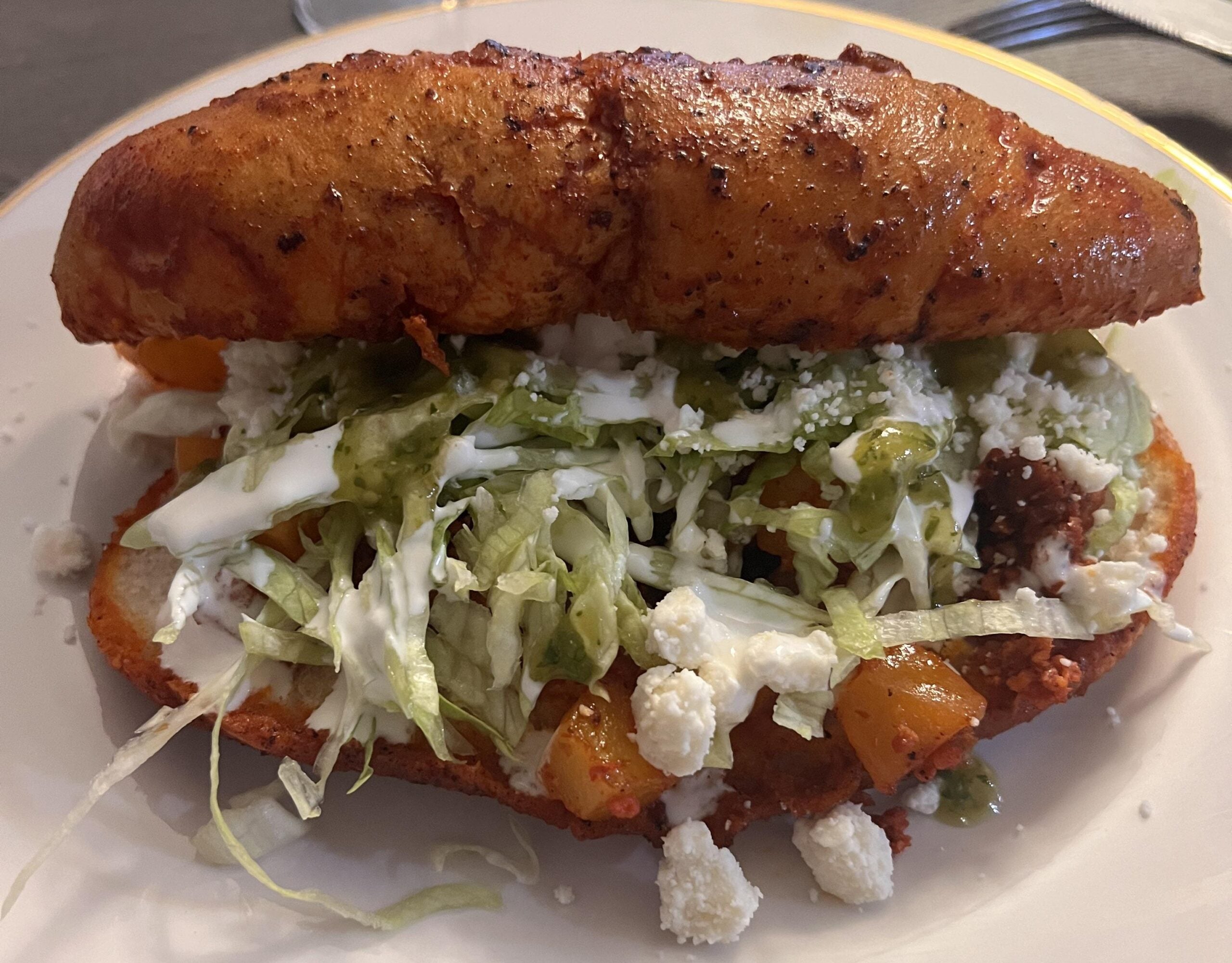 A Mexican pambazo sandwich filled with chorizo, potatoes, shredded lettuce, sour cream, green salsa, and crumbled cheese sits on a white plate.