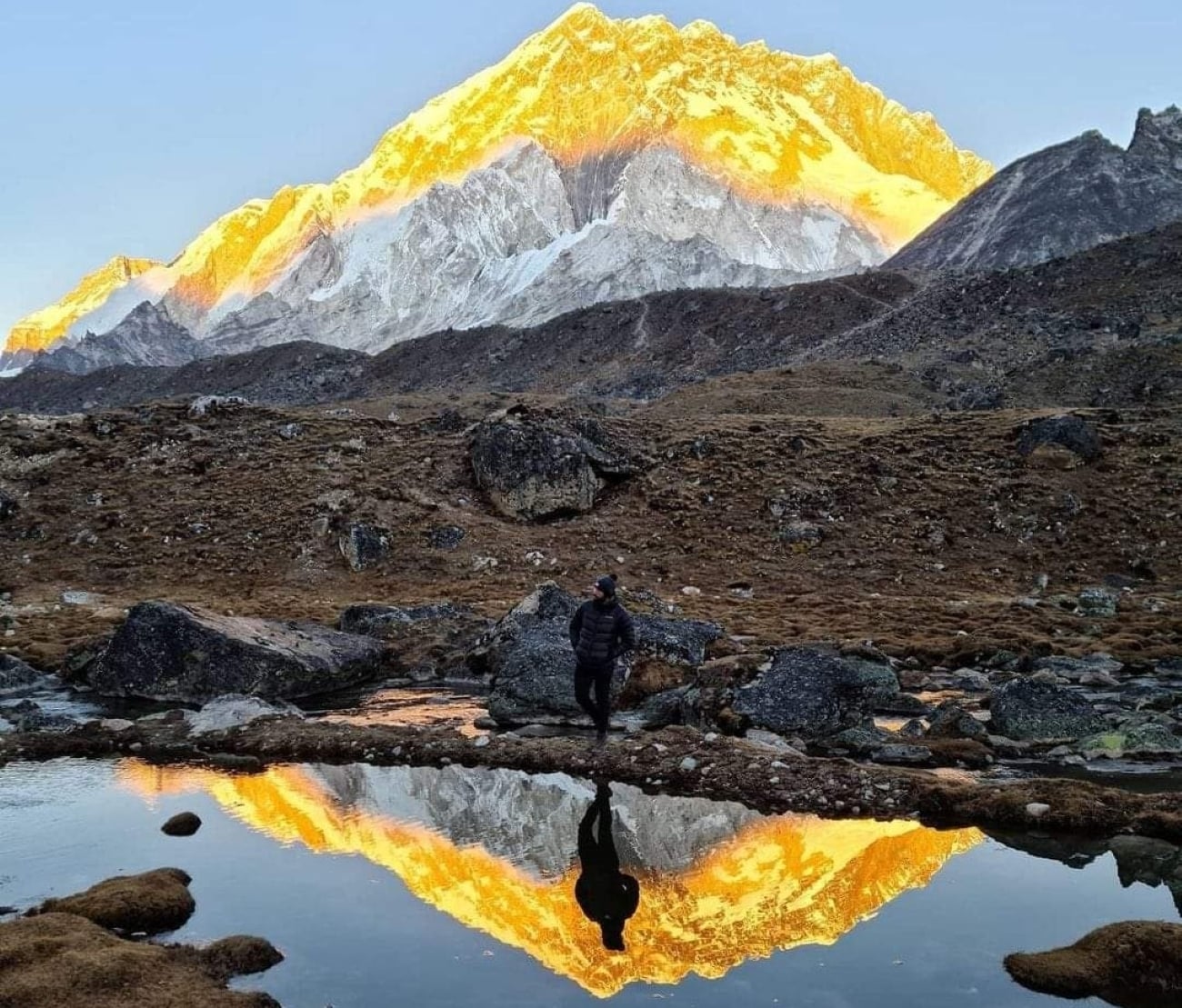 A hiker stands by a pond in a rocky landscape, with snow-covered mountains glowing in golden sunlight and their reflection visible in the water, capturing photos of the breathtaking scene to be posted later.