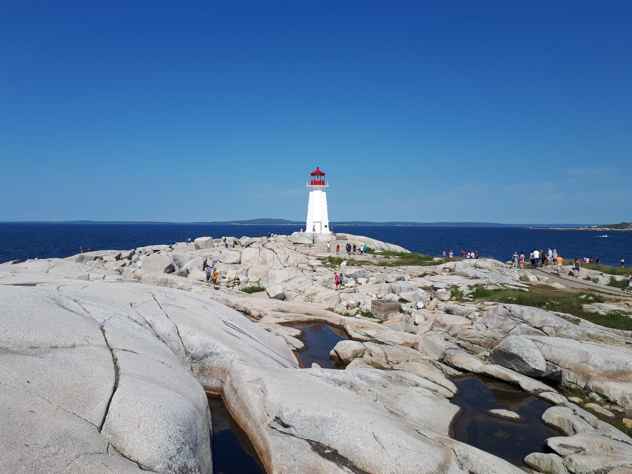 A white lighthouse with a red top stands on rocky terrain by the ocean under a clear blue sky. People are walking around the rocks and lighthouse, enjoying the sunny day.