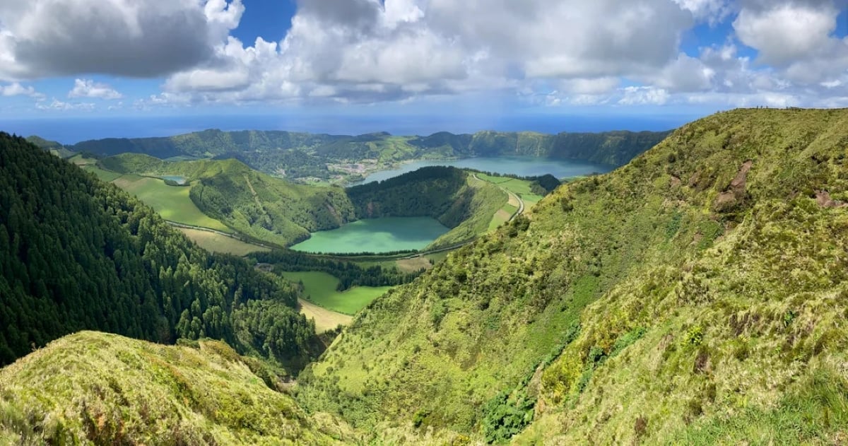 A panoramic view of lush green hills, a winding road, and lakes surrounded by forests under a partly cloudy sky.