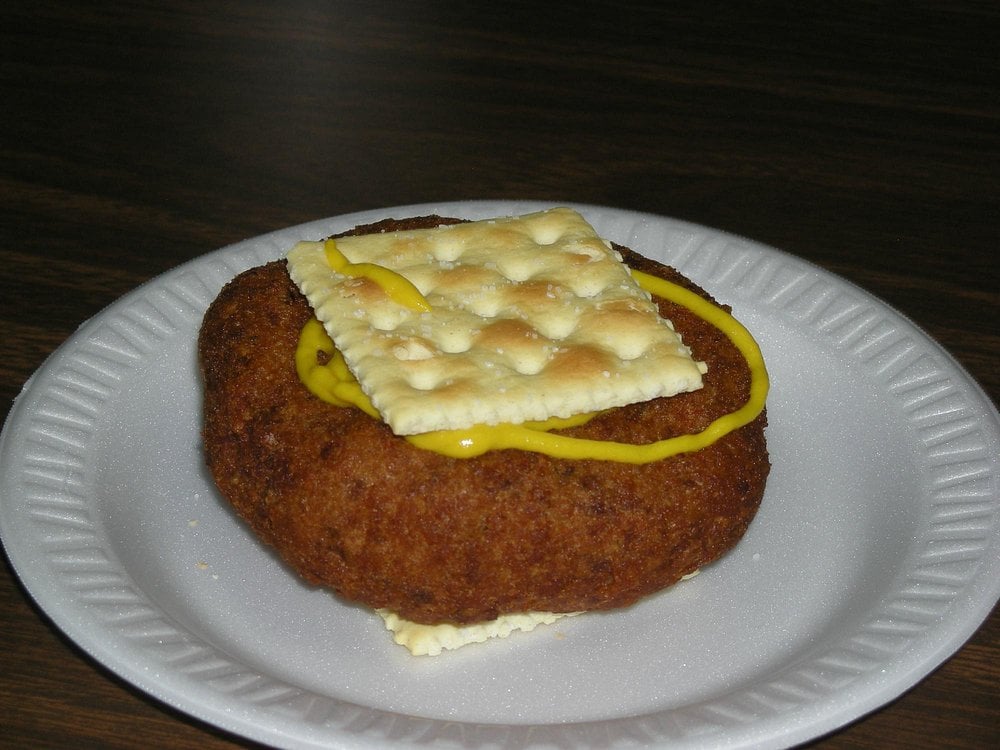 A breaded meat patty is sandwiched between two saltine crackers, with yellow mustard on top, served on a white paper plate.