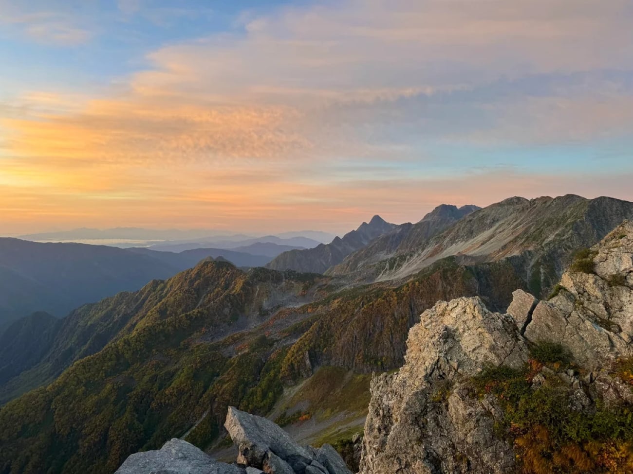 Rocky mountain ridges under a colorful sunset sky, with warm orange and pink hues reflecting on scattered clouds. Layers of distant peaks and green valleys are visible in the background, creating a breathtaking scene often posted by hikers in their photos.