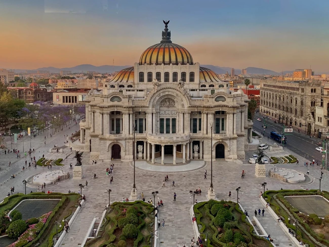 Palacio de Bellas Artes in Mexico City at sunset, with people walking in the plaza, landscaped gardens in the foreground, and city buildings in the background—a stunning scene in one of the Biggest Cities In The World.