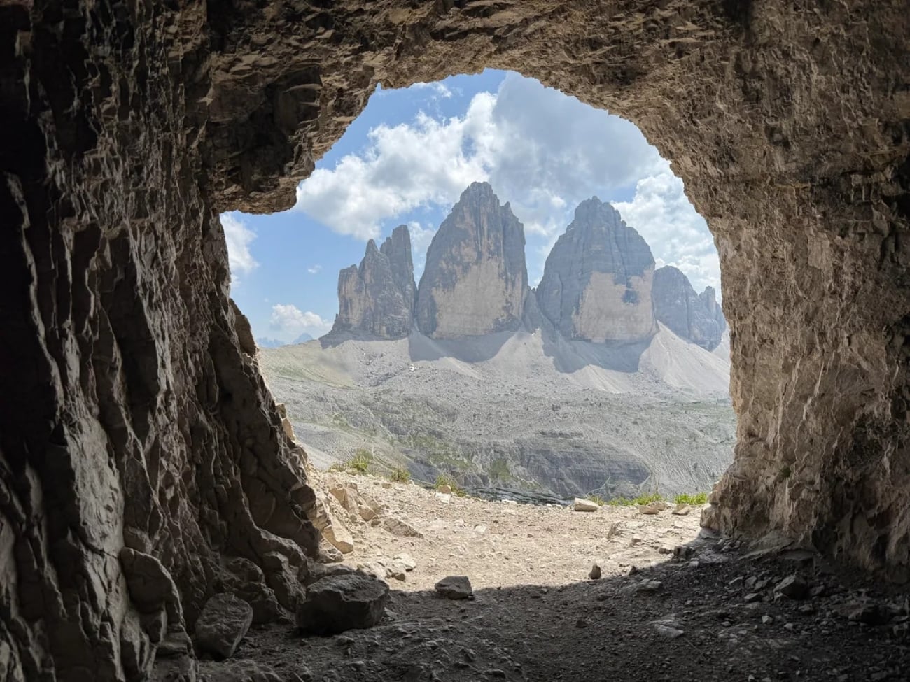 View from inside a rocky cave, looking out toward three jagged mountain peaks under a blue sky with clouds. Sunlight illuminates the mountains and the rocky ground at the cave’s entrance, creating an inviting scene often captured in hikers’ photos posted online.