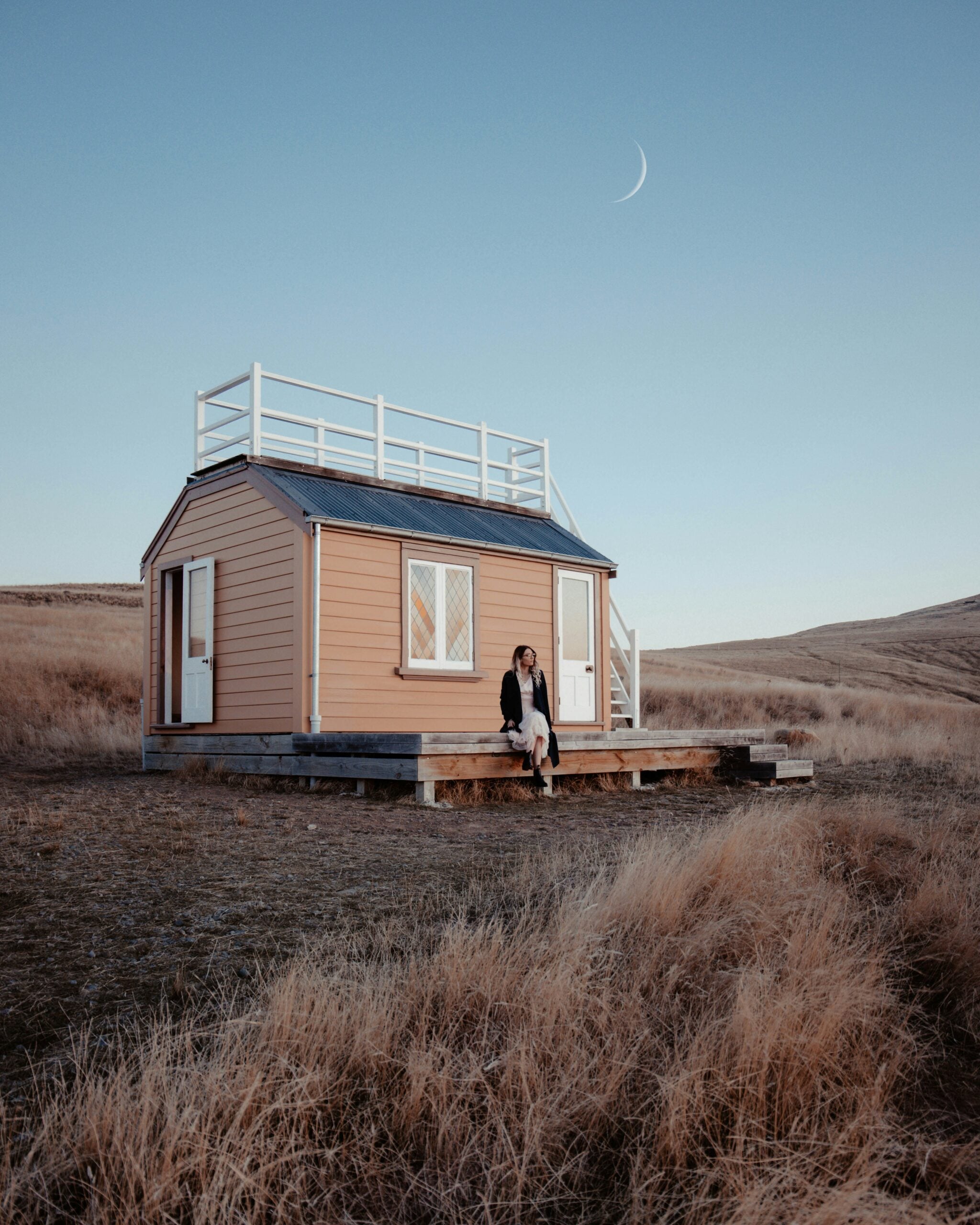 A small tan cabin stands alone in a dry, grassy field at dusk. A person sits on the front steps, and a crescent moon is visible in the clear sky above.