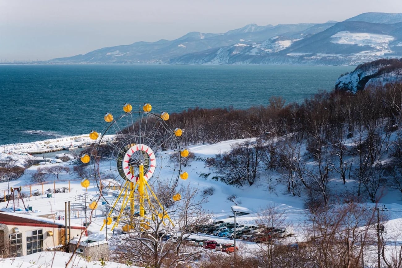 A yellow Ferris wheel stands in a snowy landscape near the sea, with a parking lot and leafless trees in the foreground and mountains in the background under a partly cloudy sky.