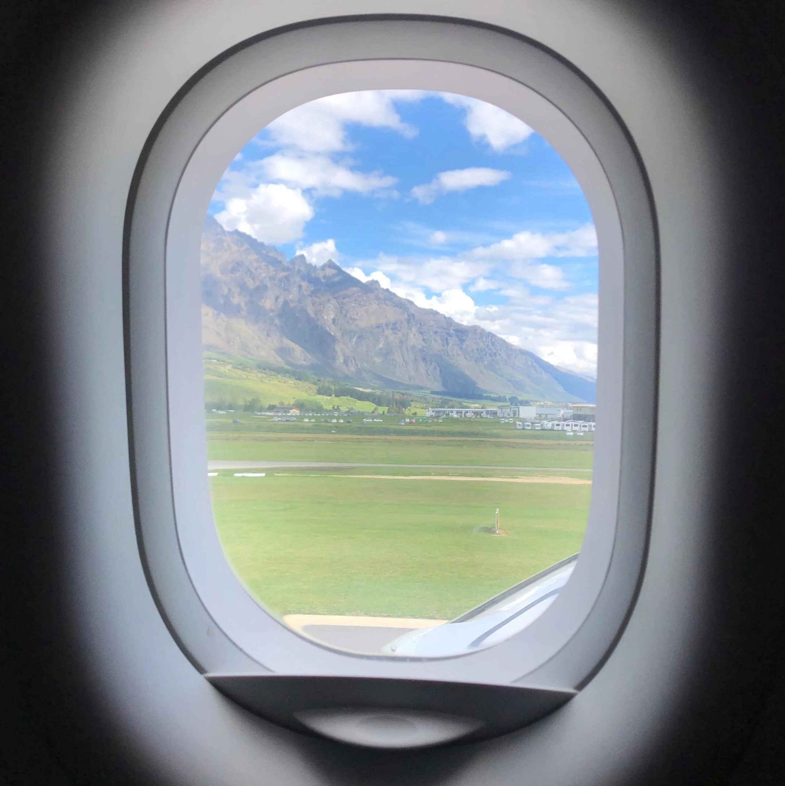 View through airplane windows showing green grass, distant mountains, blue sky, and scattered clouds. Part of a plane wing is visible at the bottom right of the window.