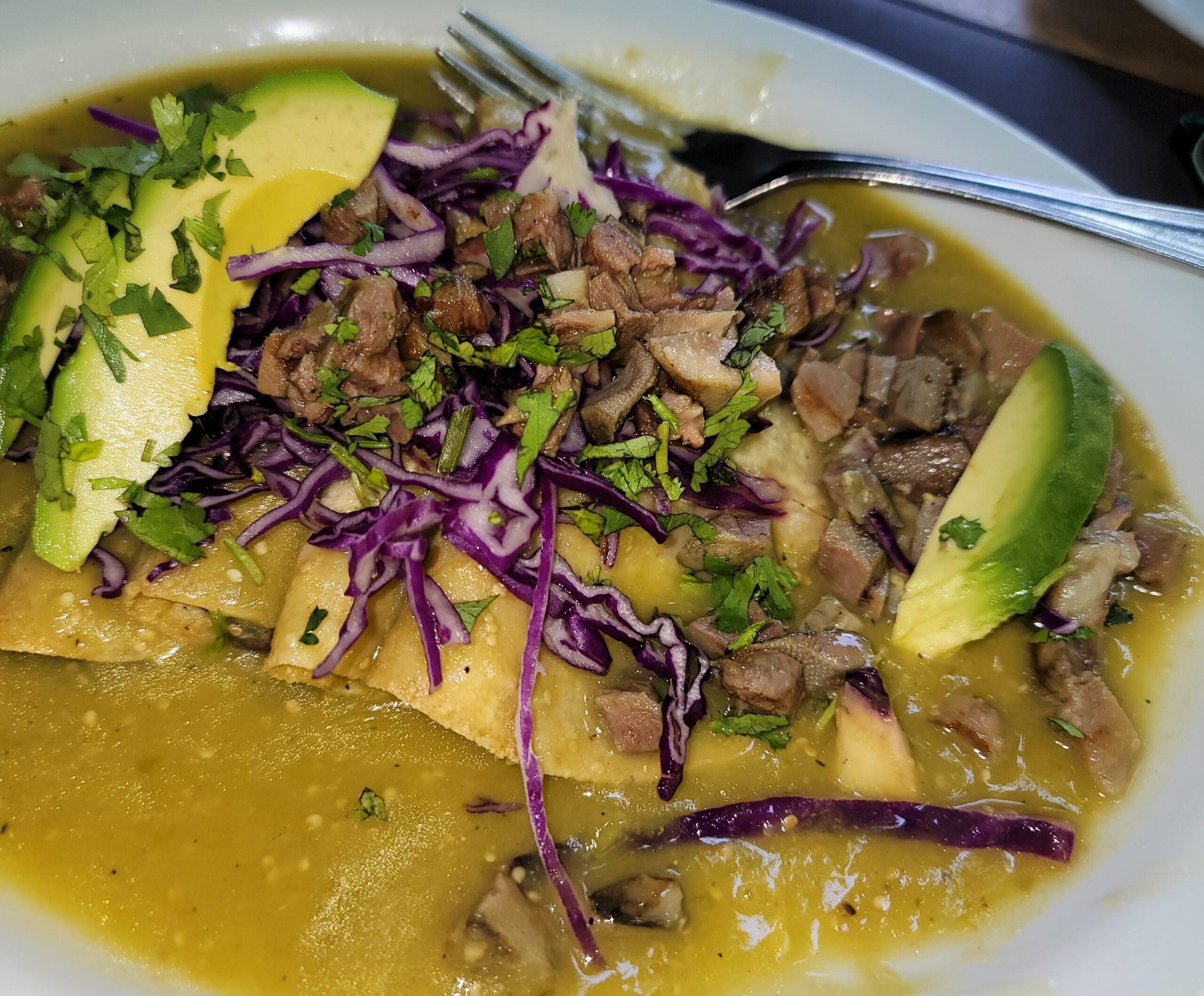 A plate of enchiladas topped with chopped beef, avocado slices, shredded purple cabbage, cilantro, and green sauce, with a fork on the side.