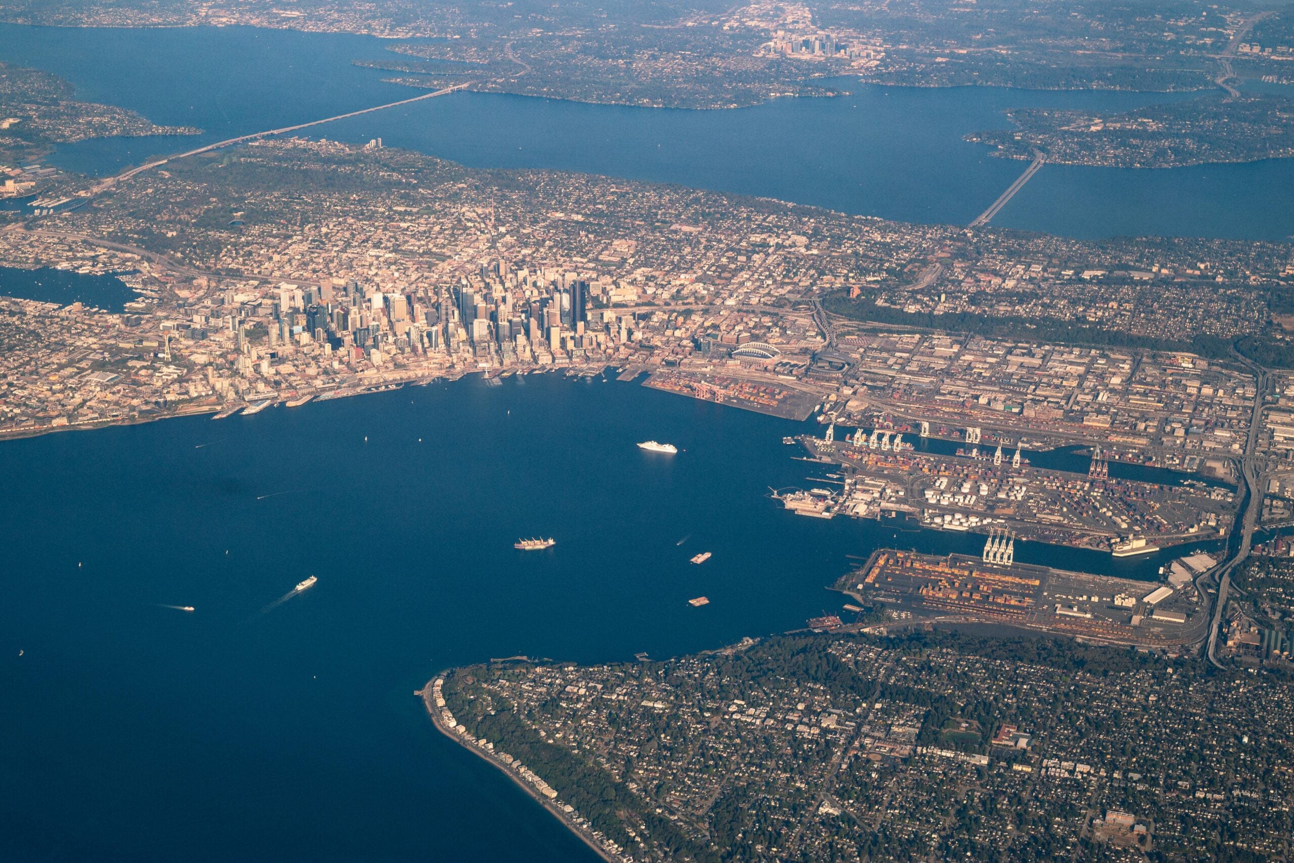 Aerial view of a coastal city with skyscrapers by the water, seen as if looking through airplane windows, showcasing a busy harbor with ships and bridges connecting landmasses, all surrounded by vibrant urban and green areas.