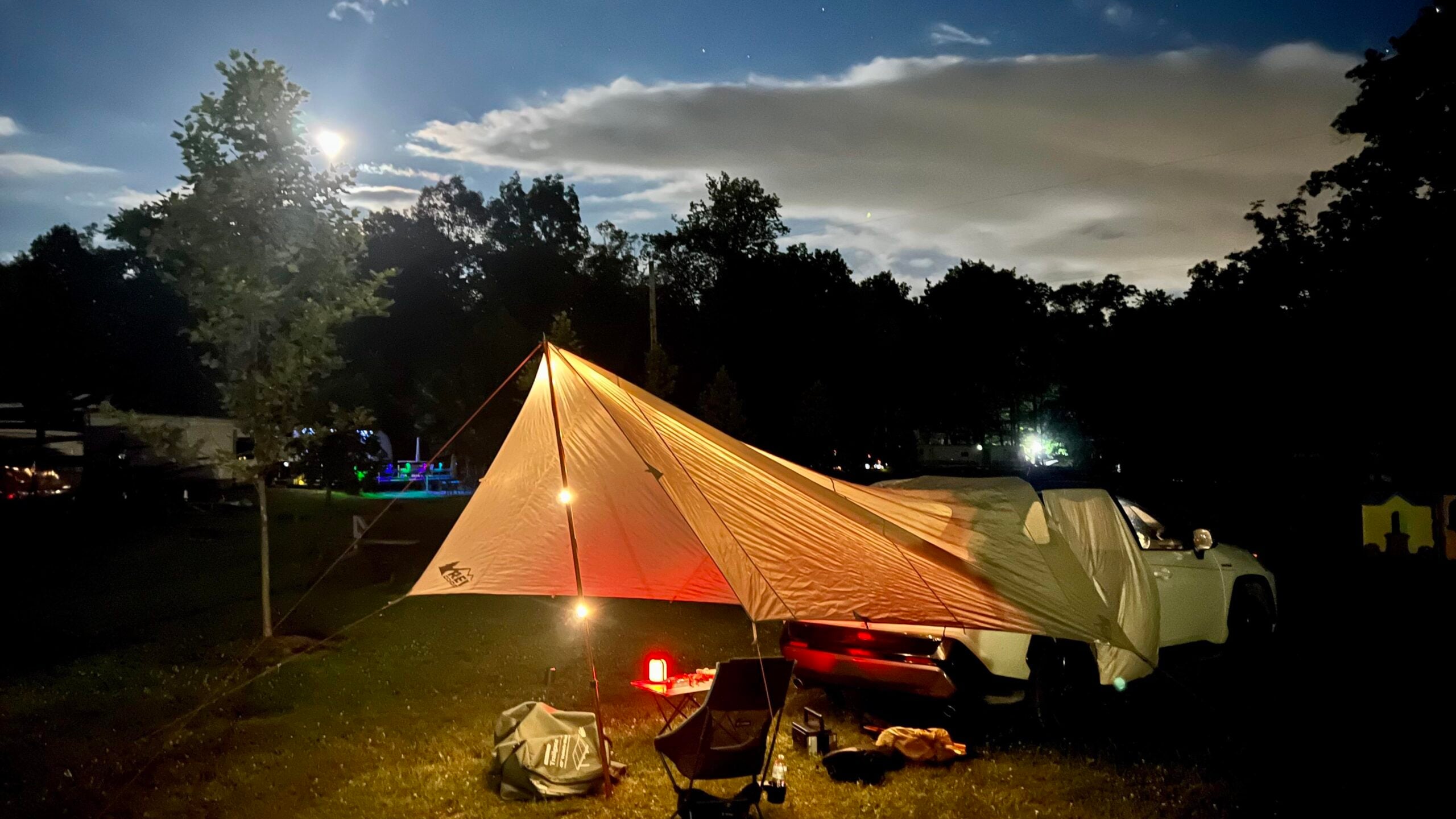 A nighttime campsite with a white pickup truck and a large tarp lit by lanterns, set up near trees under a bright moon and cloudy sky. Camping chairs and gear are visible under the tarp.
