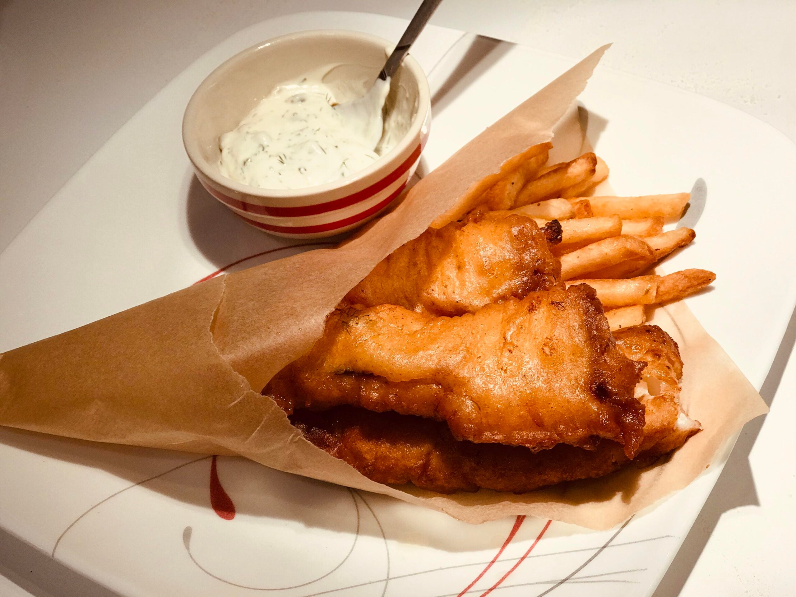 Golden fried fish fillets and French fries in brown paper, served on a plate with a small bowl of creamy dip and a spoon.
