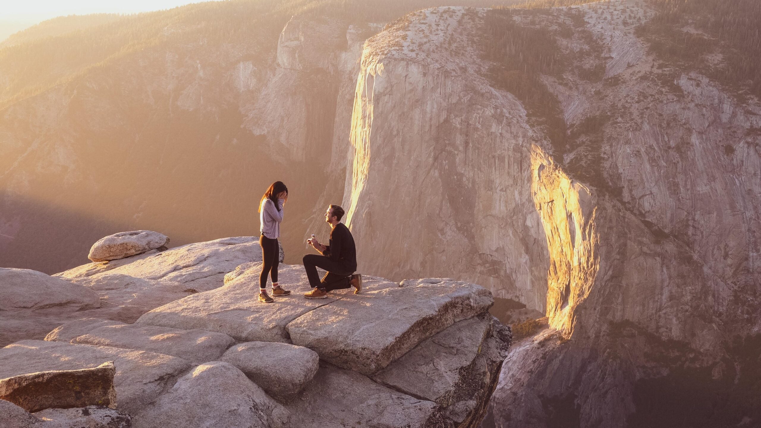 A person kneels and proposes to another on a rocky cliff at sunset, with dramatic mountains and warm sunlight in the background—capturing one of those unforgettable moments often posted by hikers in their adventure photos.