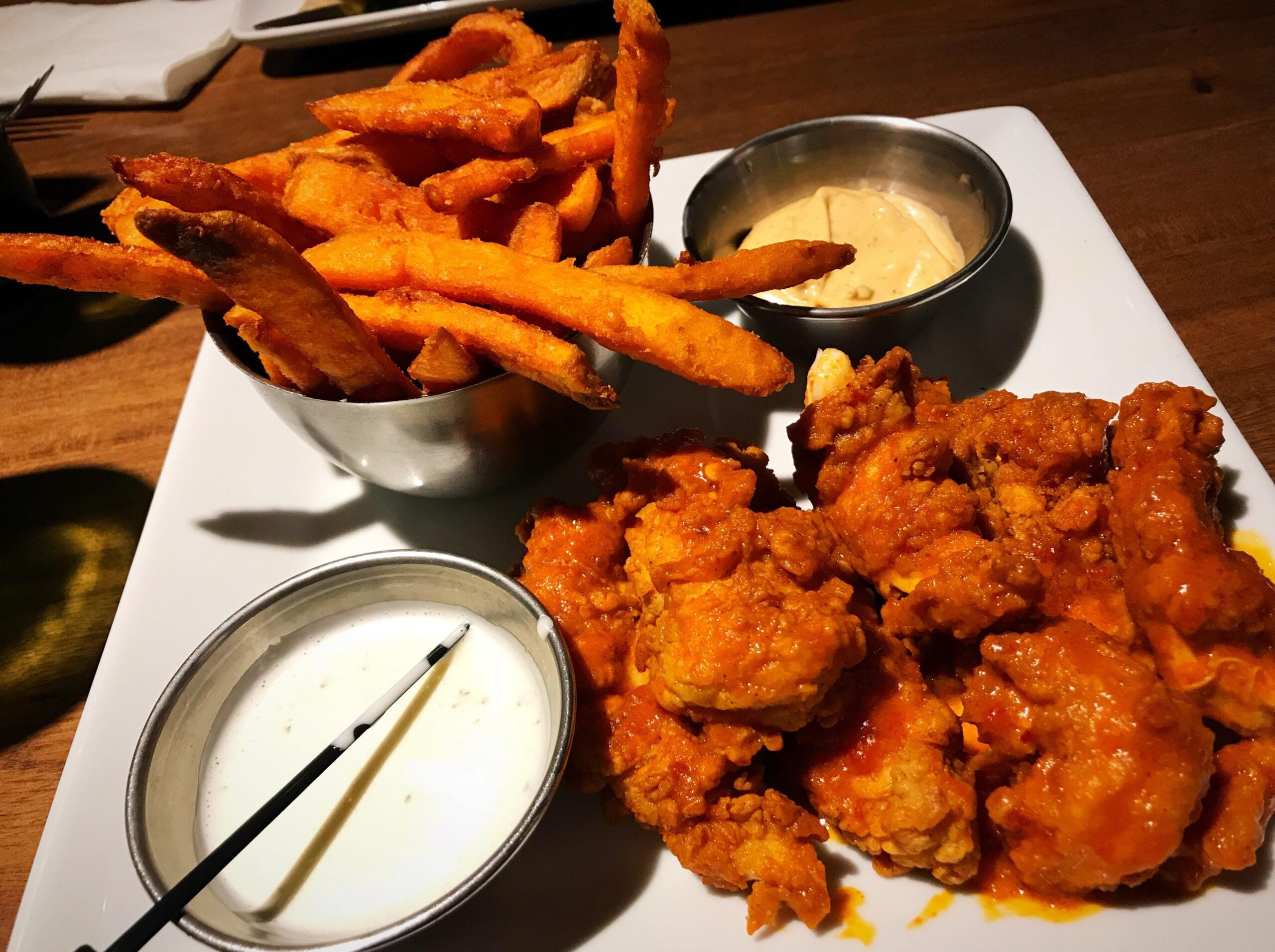 A plate with buffalo chicken bites, a cup of dipping sauce, sweet potato fries in a metal bowl, and another dipping sauce on a white square plate, set on a wooden table.