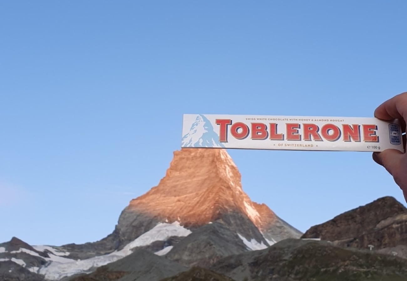 A hand holds a Toblerone chocolate bar so it aligns with the peak of a mountain in the background, perfectly mimicking photos hikers posted where the mountain resembles the chocolate bar’s iconic peak on the packaging.