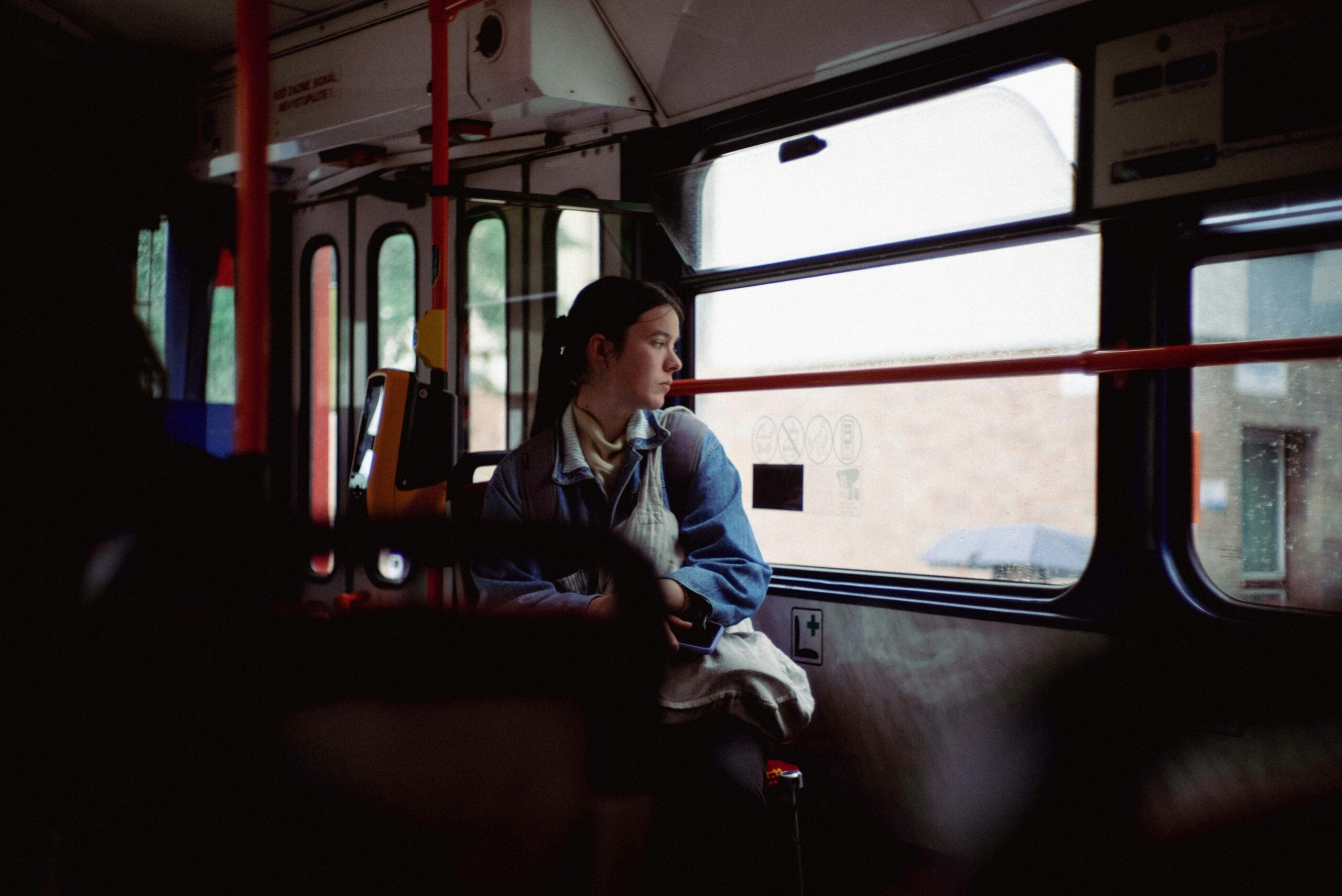 A person wearing a denim jacket sits alone on a bus, looking out the window with a thoughtful expression. The outside appears rainy and the bus interior is dimly lit.