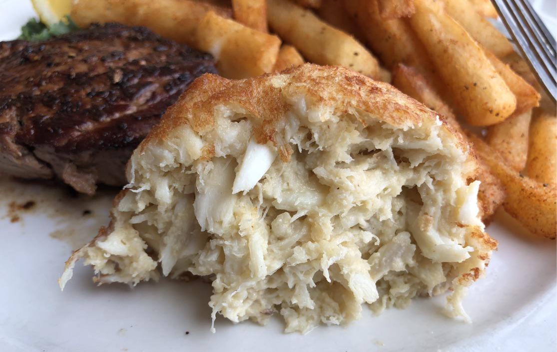 A close-up of a crab cake showing its flaky texture, with seasoned steak fries and part of a grilled steak on a white plate in the background.
