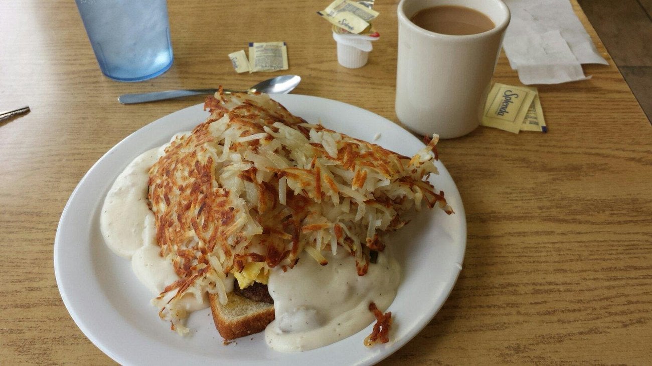 A breakfast plate with hash browns, white gravy, and toast sits on a wooden table next to a mug of coffee, a glass of water, cream, and sugar packets.