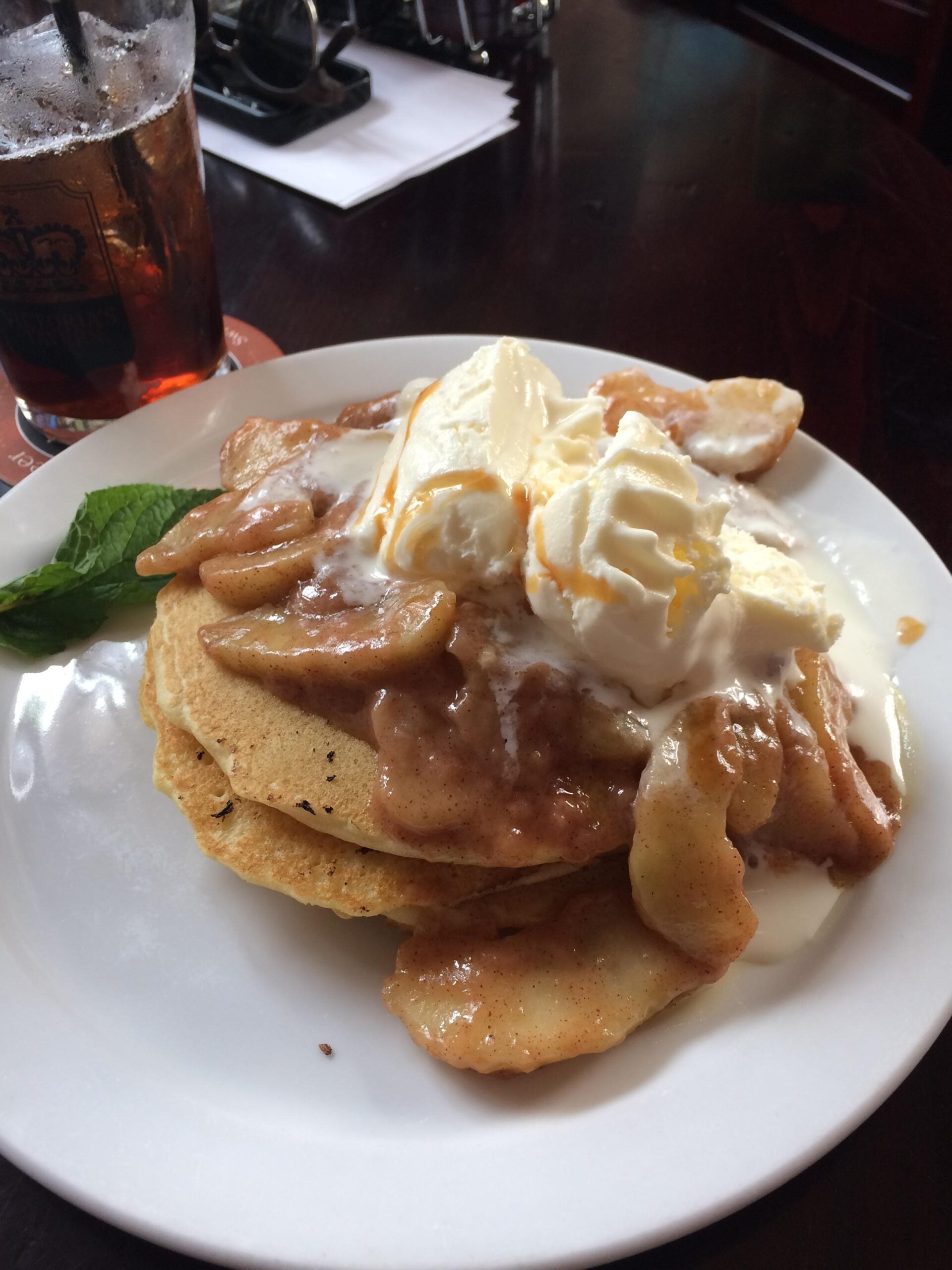 A plate of pancakes topped with caramelized apple slices and melting vanilla ice cream, garnished with a mint leaf. A glass of soda and some papers are in the background on a dark table.