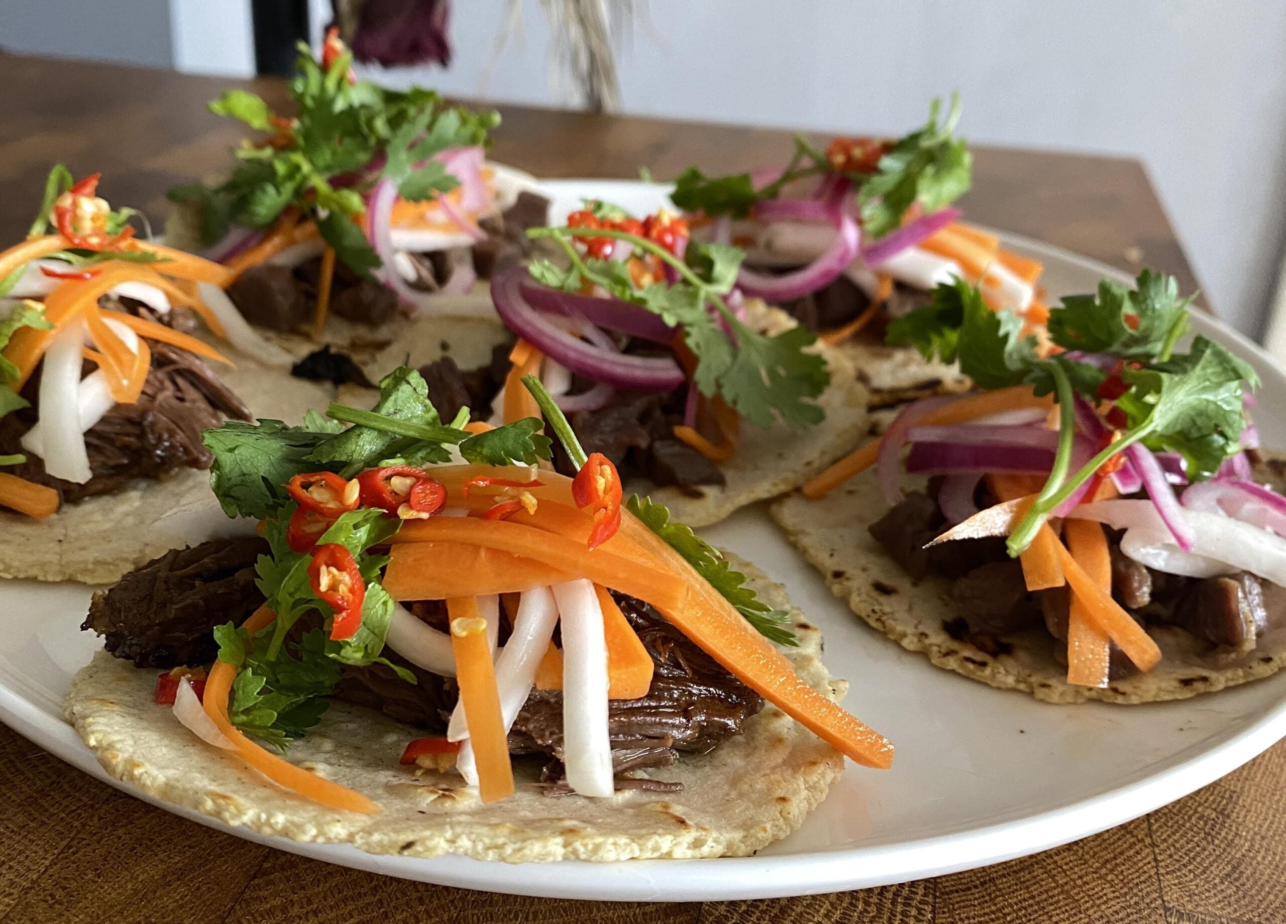 Plate of tacos topped with shredded beef, pickled carrots, daikon, red onions, sliced red chili, and fresh cilantro on corn tortillas, arranged neatly on a white plate.