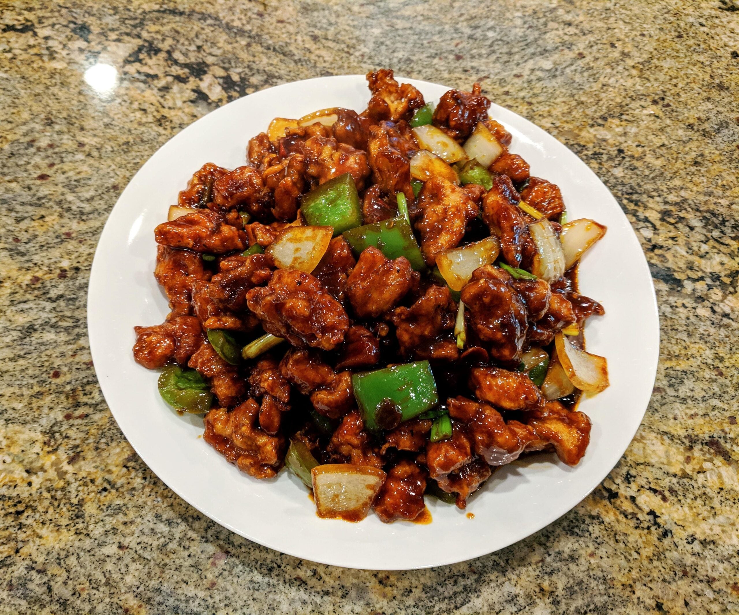 A white plate filled with stir-fried chicken, green bell peppers, and onions coated in a dark sauce sits on a granite countertop.