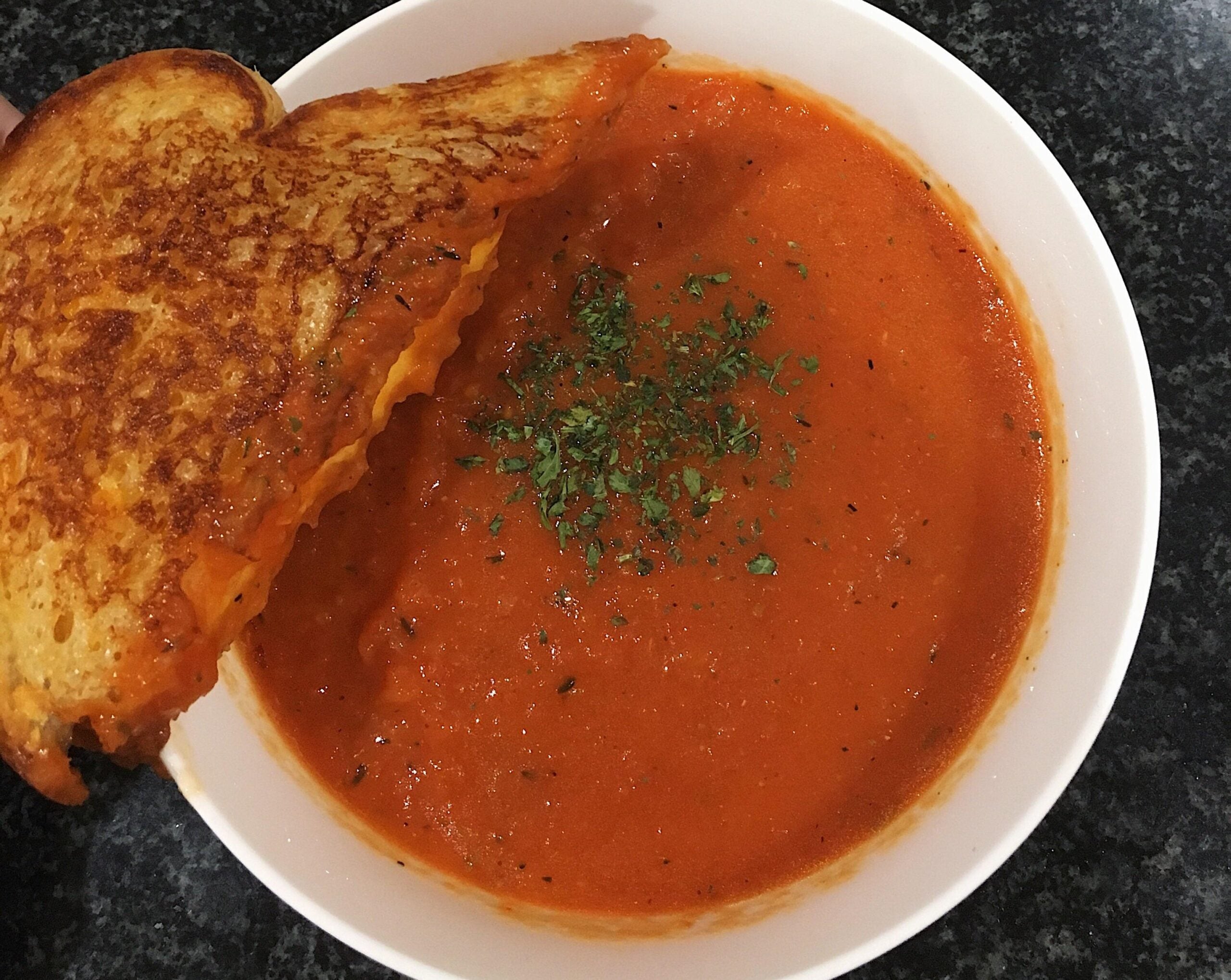 A bowl of tomato soup garnished with herbs, with a grilled cheese sandwich partially dipped into the soup.
