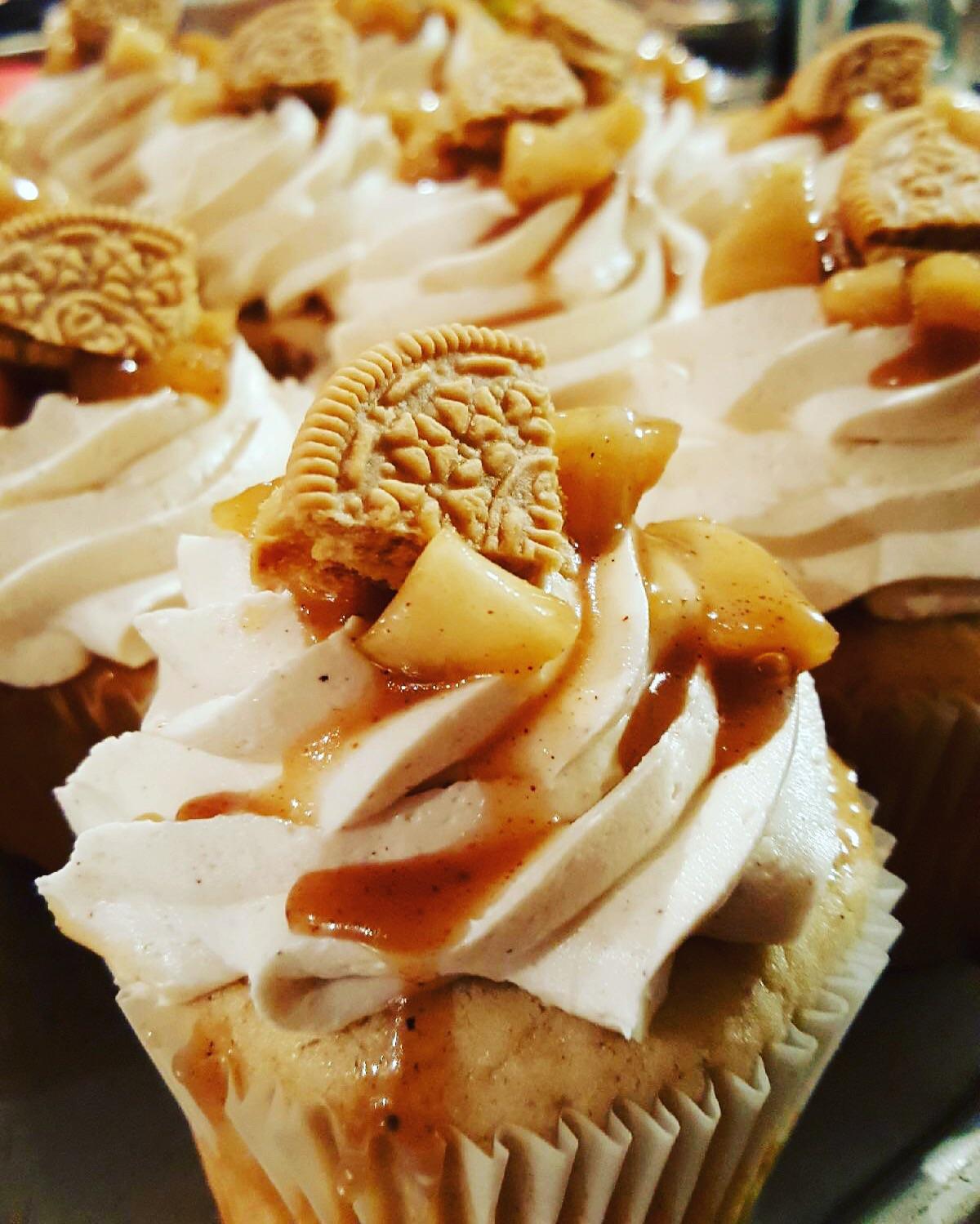 Cupcakes topped with swirls of frosting, cookie pieces, diced apples, and caramel sauce, arranged closely together.